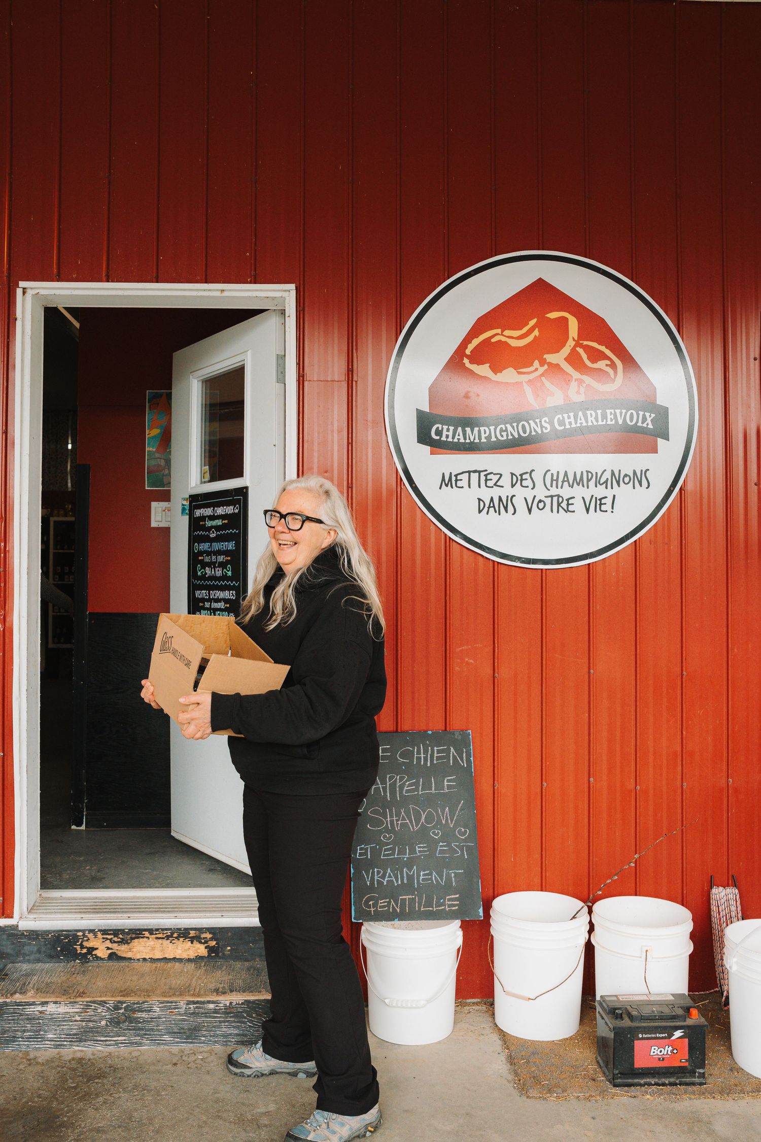 Woman holding a cardboard box outside Champignons Charlevoix mushroom farm red building.