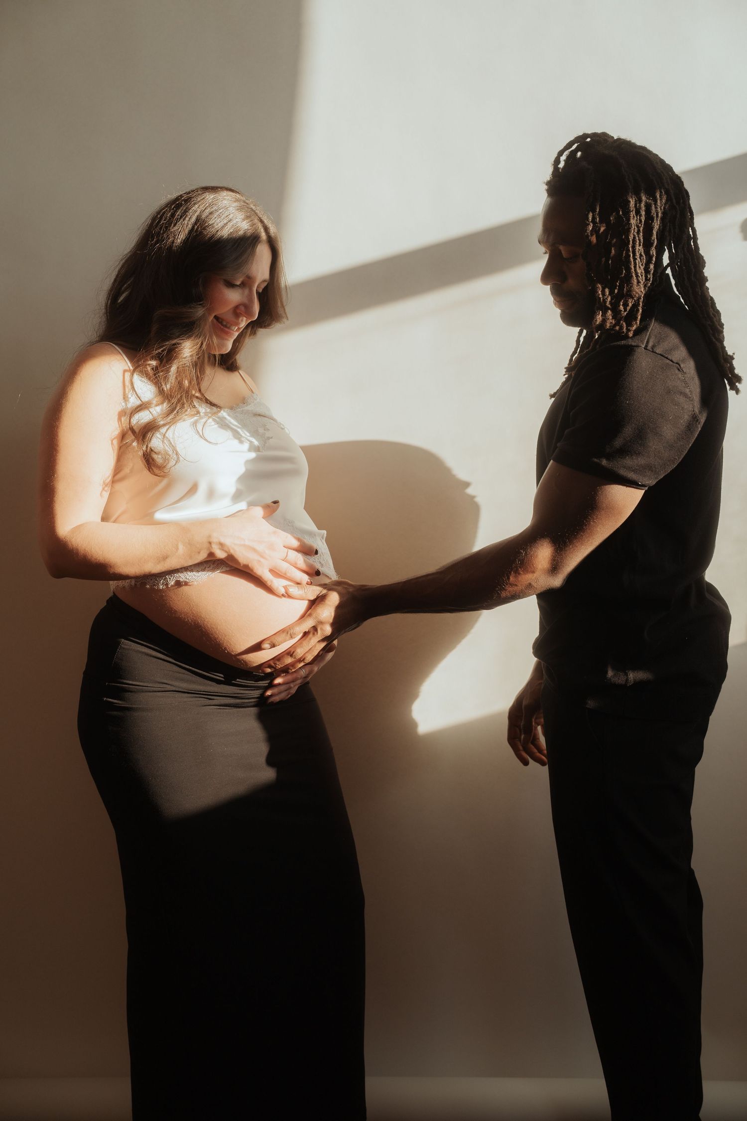 A pregnant woman in a white top and black skirt cradles her baby bump as her partner tenderly touches her belly in soft sunlight.