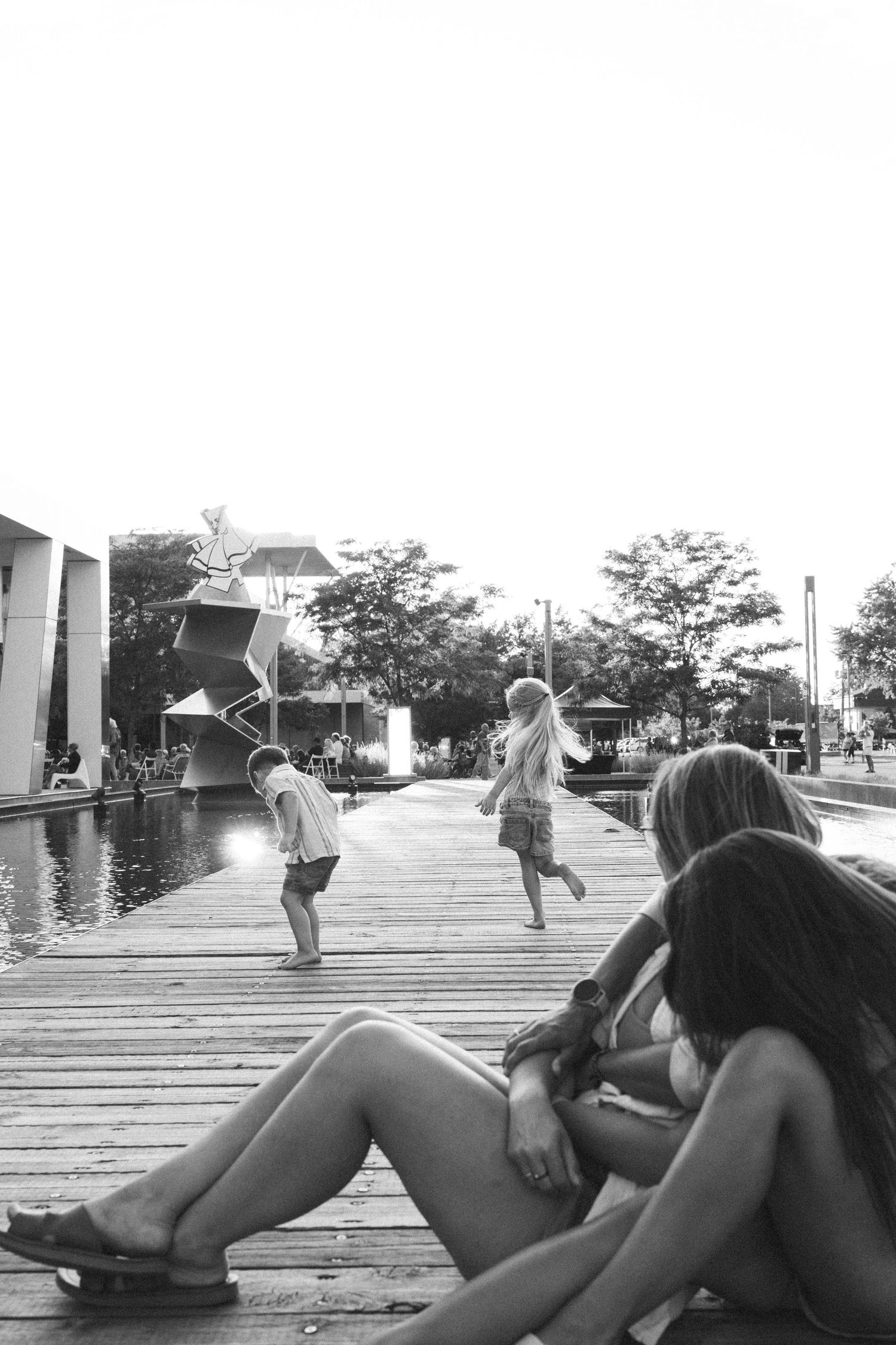 Black and white photo of children playing on a wooden boardwalk near water while two women relax in the foreground.