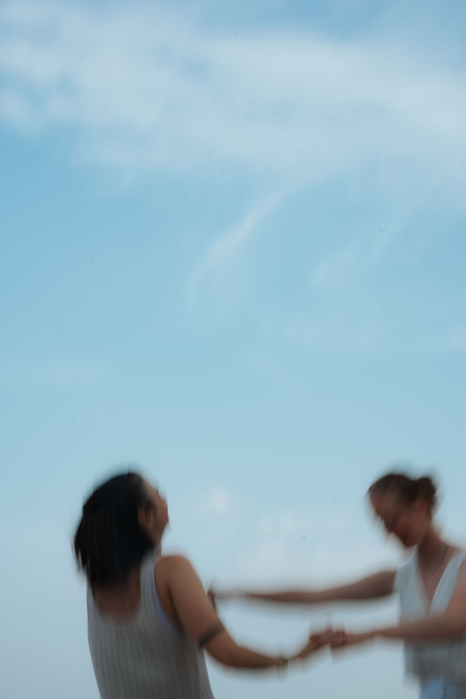 Two women in white dresses hold hands dancing under a bright blue sky in a blurred motion photo.