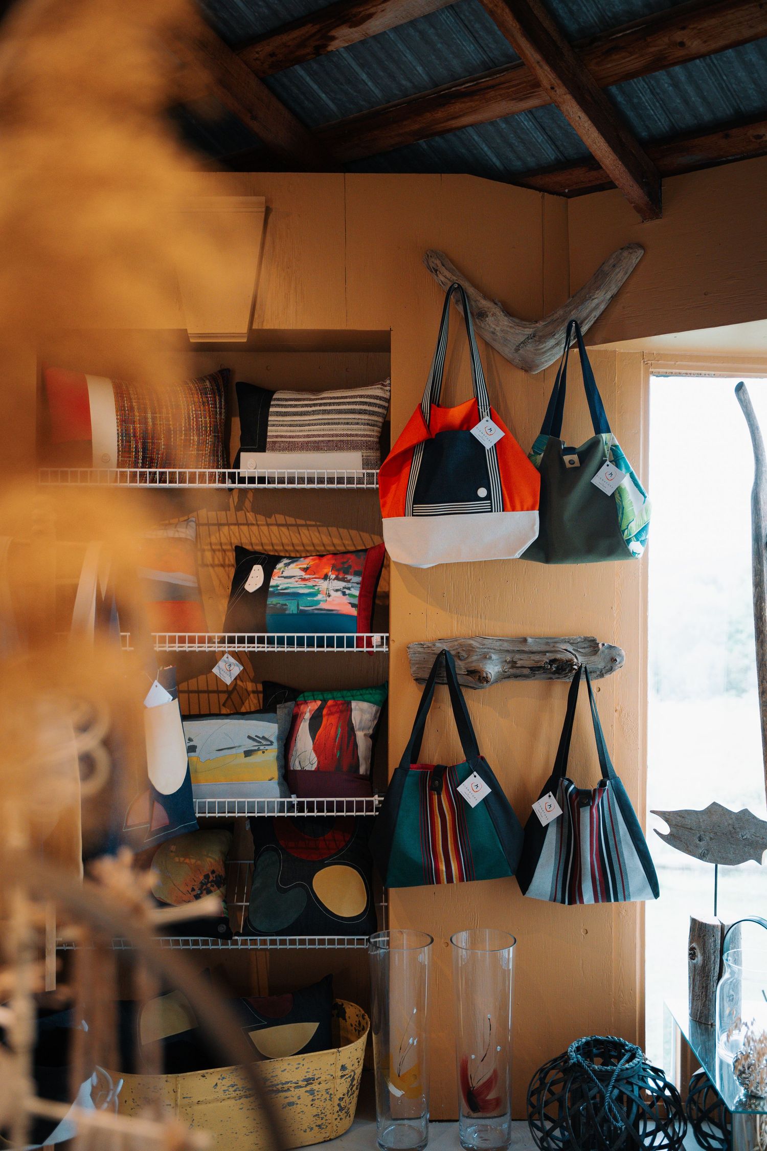Colorful tote bags hanging on driftwood display with shelves of folded textiles in a rustic boutique shop.