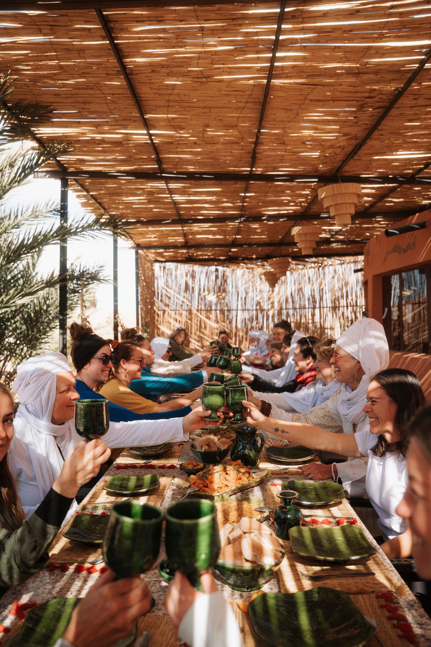 Group of people raising green glasses in a toast under a bamboo roof at a Moroccan dinner.