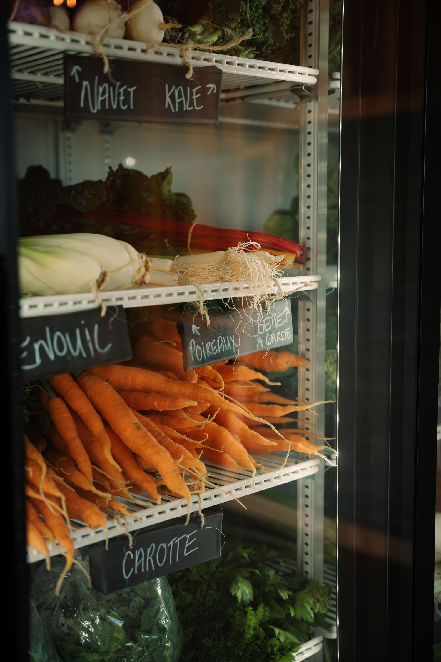 Refrigerator display of fresh vegetables including carrots, fennel, and kale with chalkboard labels in a grocery store.