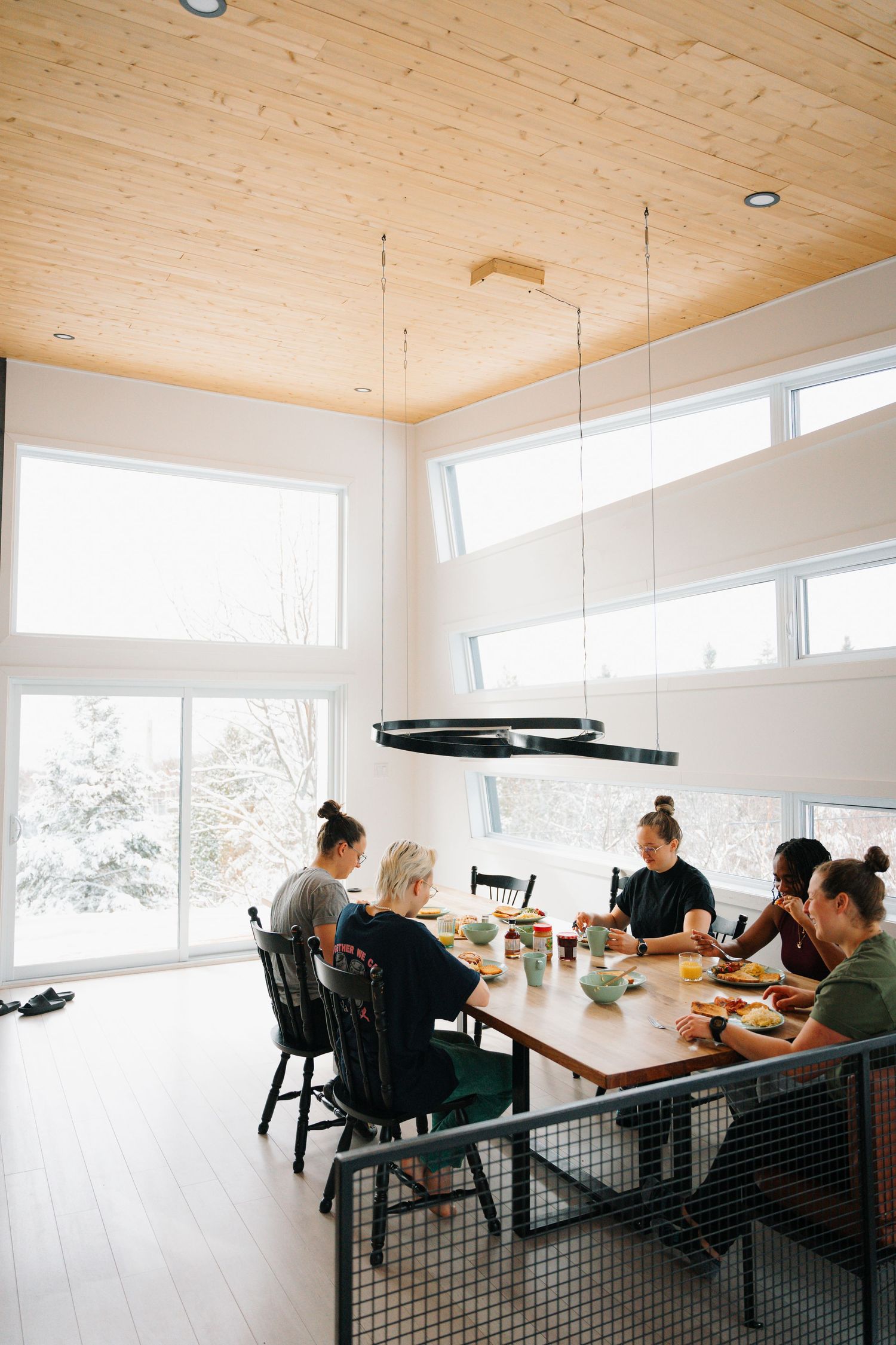 Group of people dining together at a large wooden table in a bright modern cabin interior.