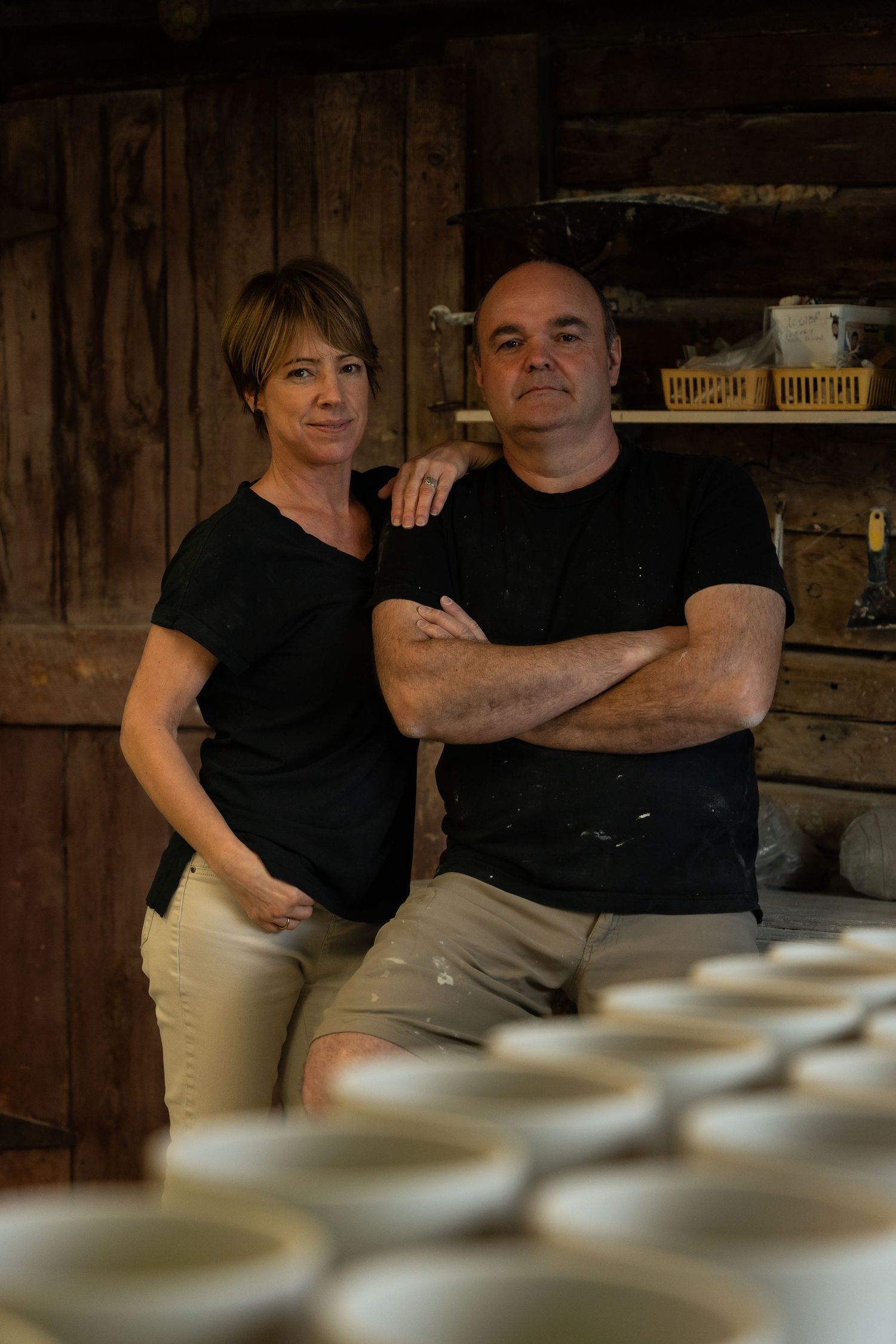 Two ceramic artists posing in their pottery studio with unfired clay bowls in the foreground.