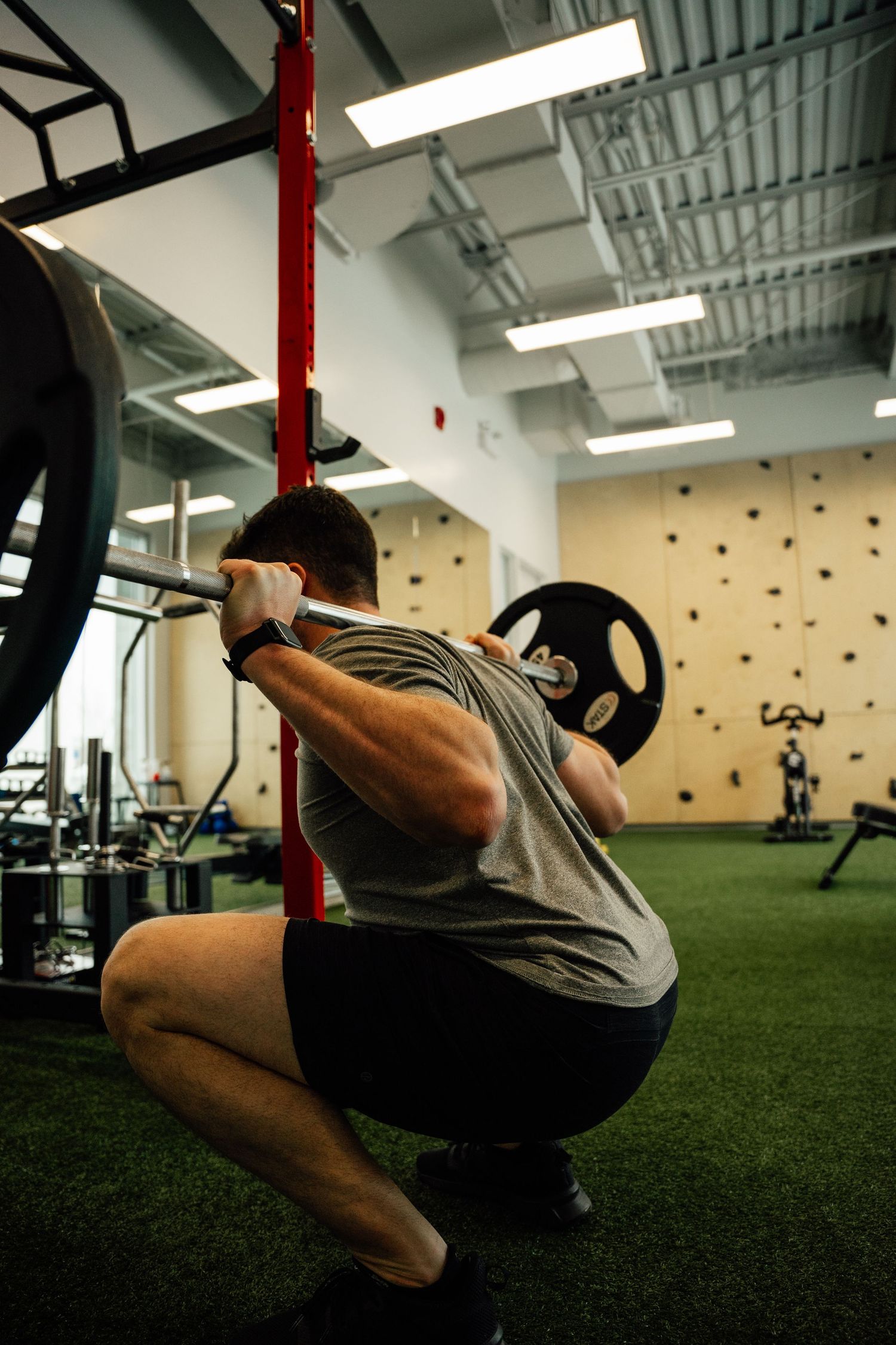Athlete performing a barbell back squat in a modern gym with a climbing wall in the background.