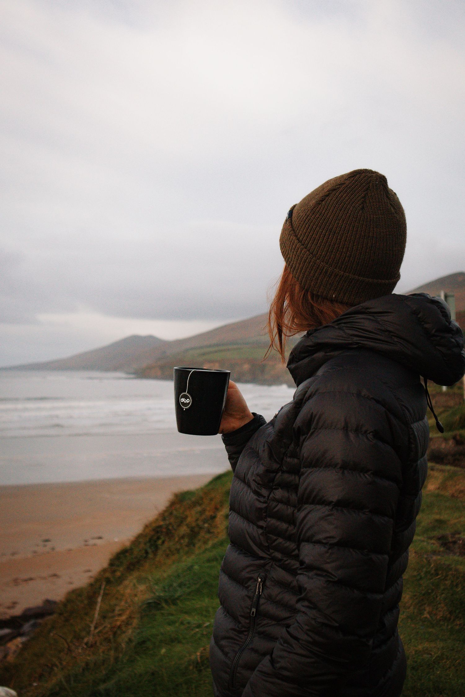 Woman in black puffer jacket and brown beanie holding mug overlooking scenic beach.