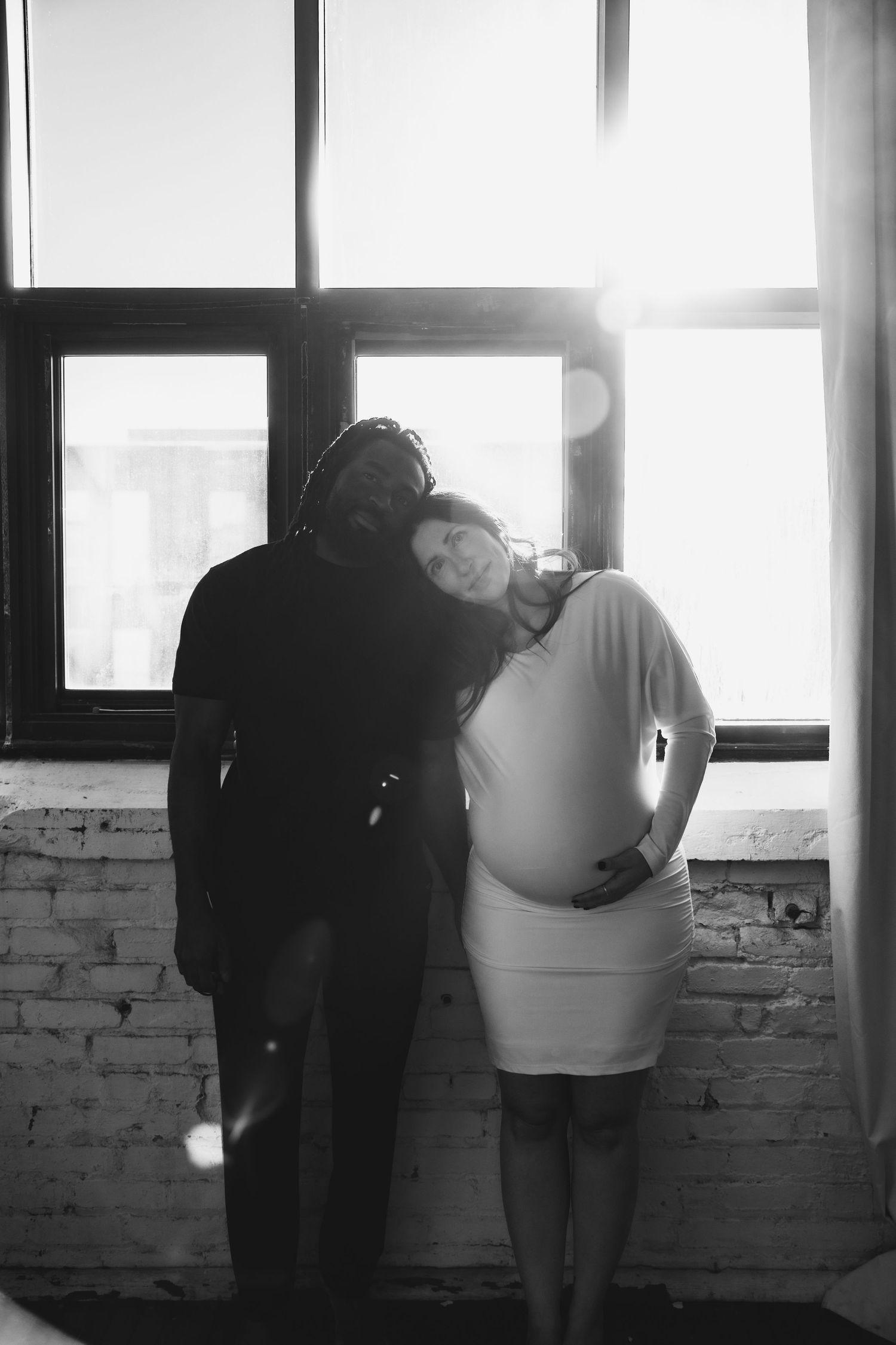 Black and white photo of interracial couple embracing by sunlit window, woman showing baby bump.