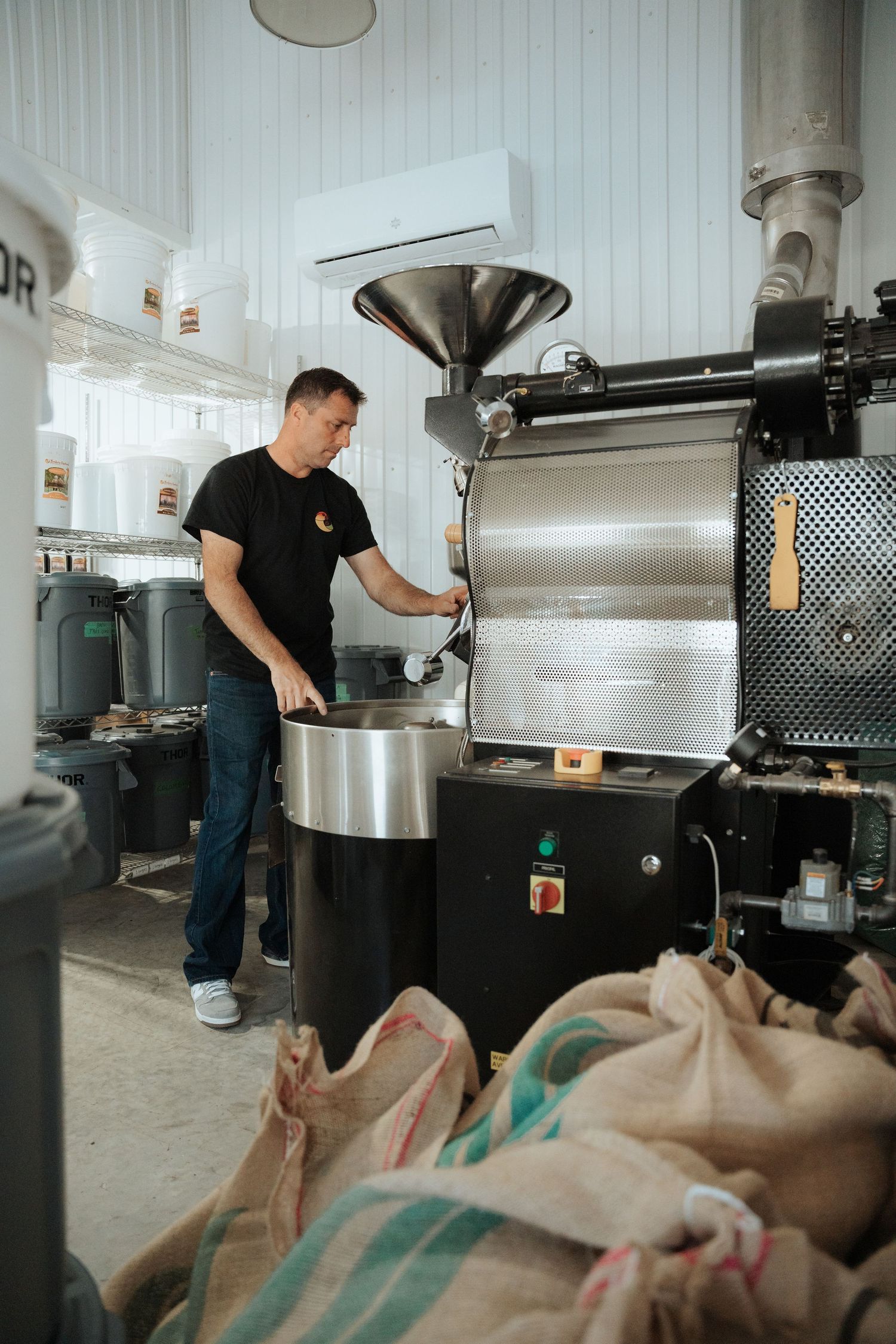 Man in black t-shirt operating a professional coffee roasting machine in an industrial roastery with burlap sacks.