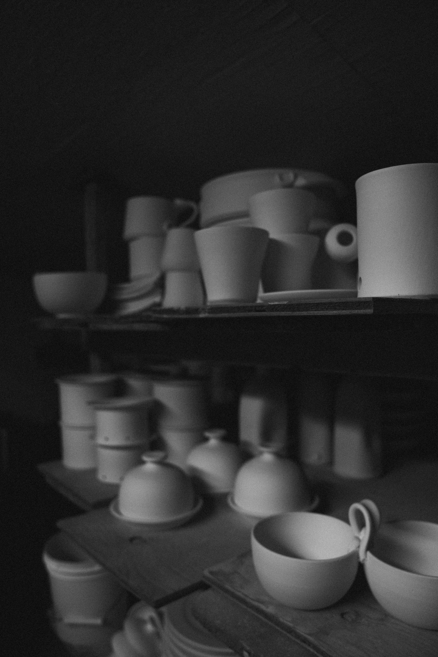 Black and white photo of unfired ceramic cups and bowls on wooden shelves in a pottery studio.