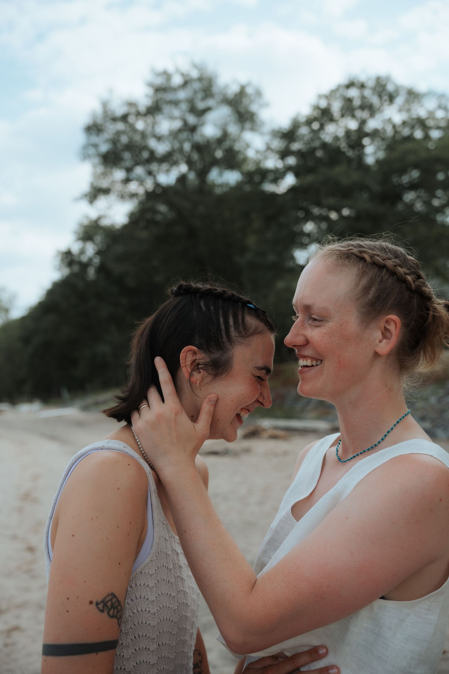 Two brides in white laugh and embrace on a sandy beach with trees in the background.