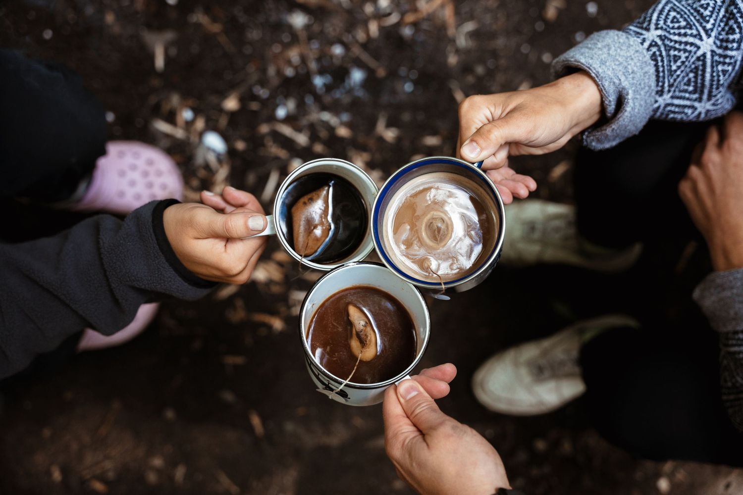 Three people toasting with enamel camping mugs filled with hot chocolate outdoors.