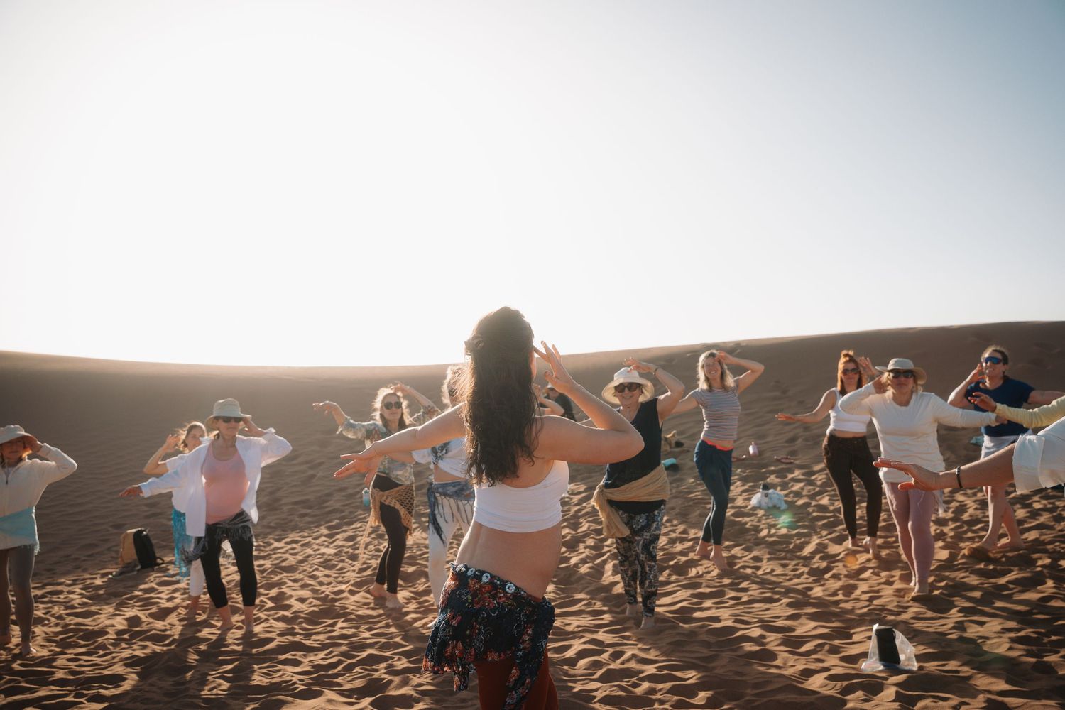 Instructor leading a group belly dance class on sandy desert dunes at golden sunset.