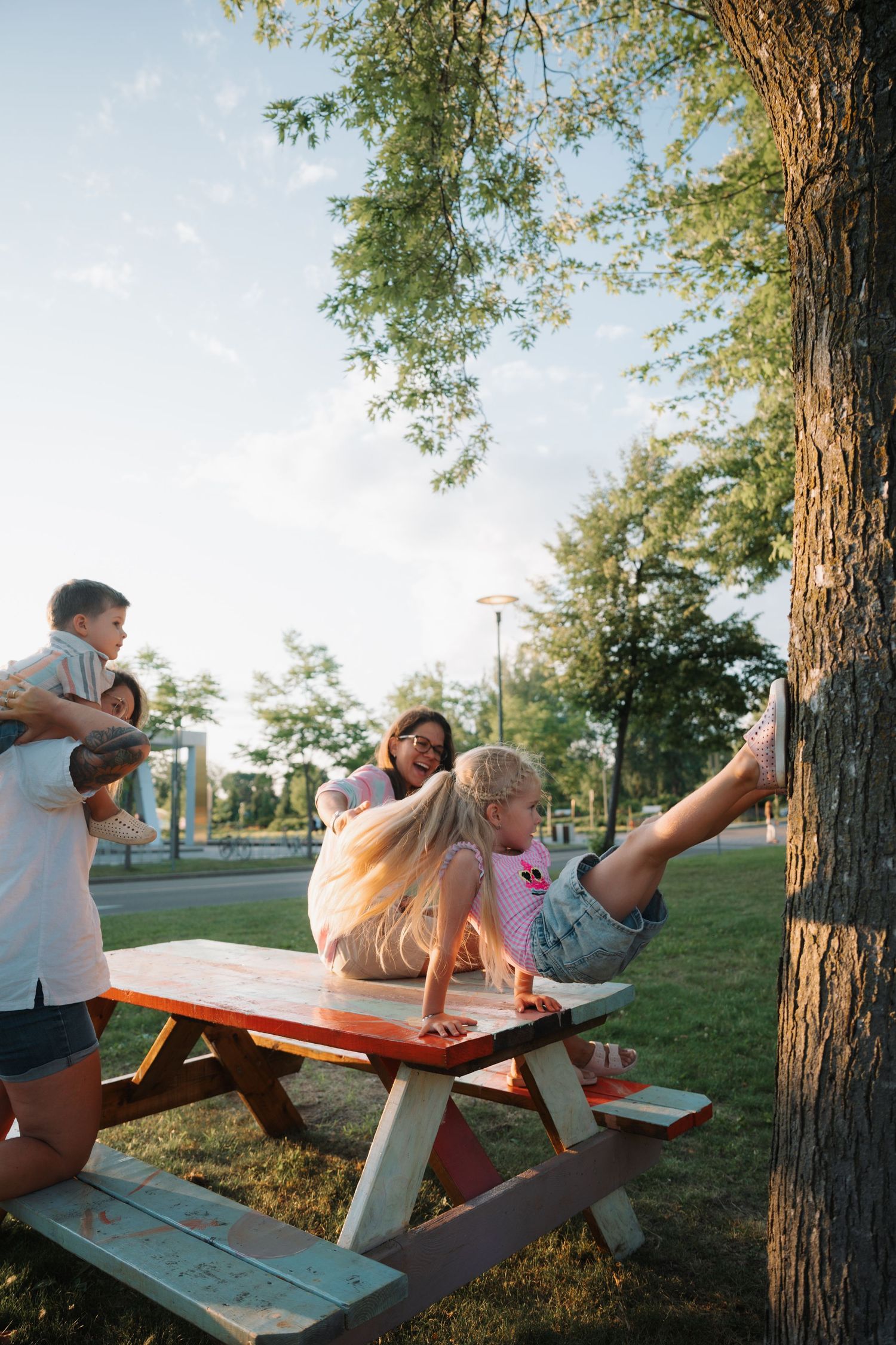 Family enjoying a sunny day at a picnic table in the park, with children playing near a large tree.