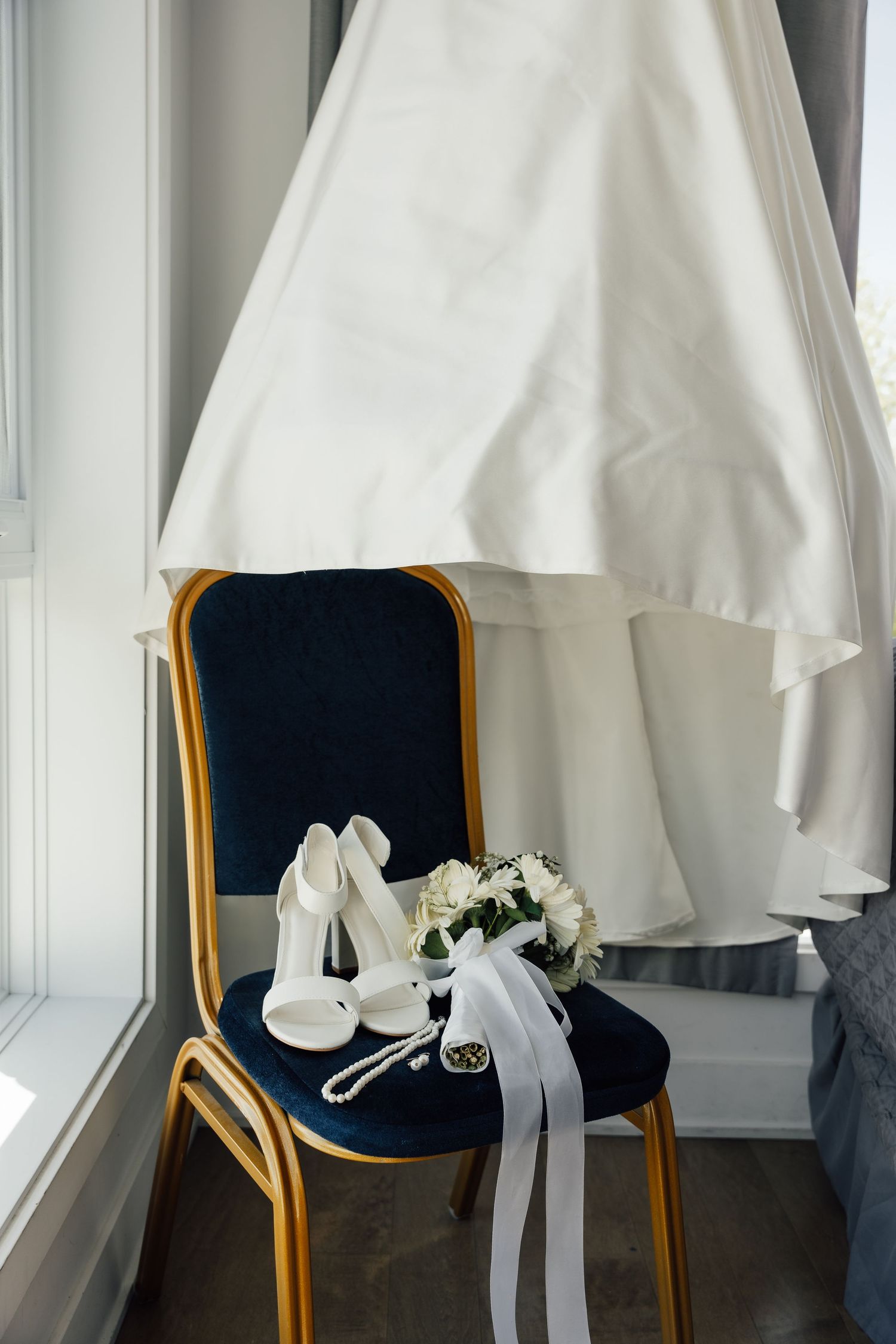 White bridal heels, a pearl necklace, and a small bouquet sit on a chair beneath a hanging wedding gown.