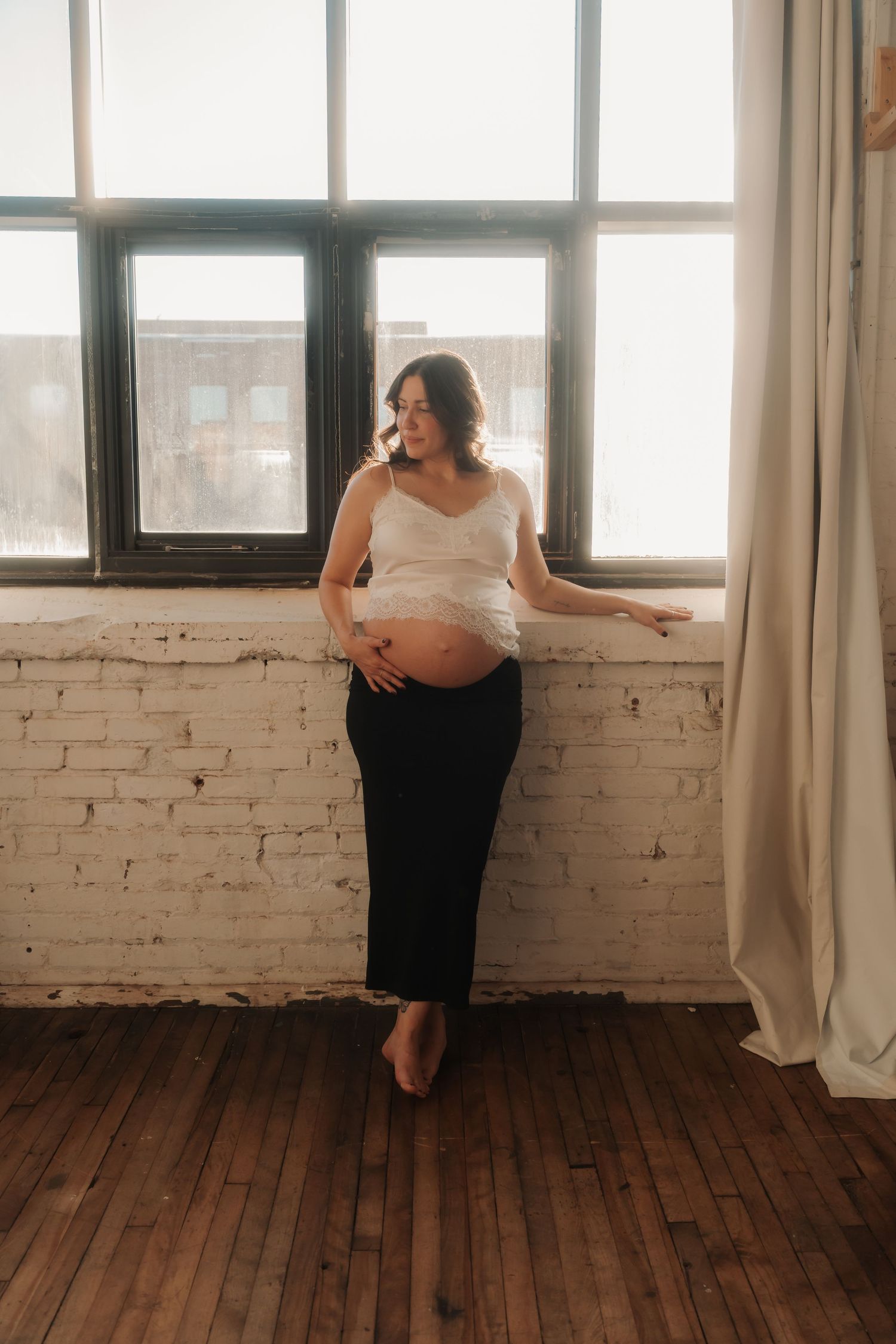 Pregnant woman in white top and black skirt leaning against brick wall by large windows in natural light.