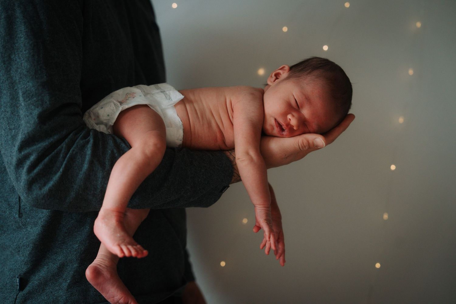 Newborn baby sleeping peacefully while being held face-down on parents arm against bokeh light background.