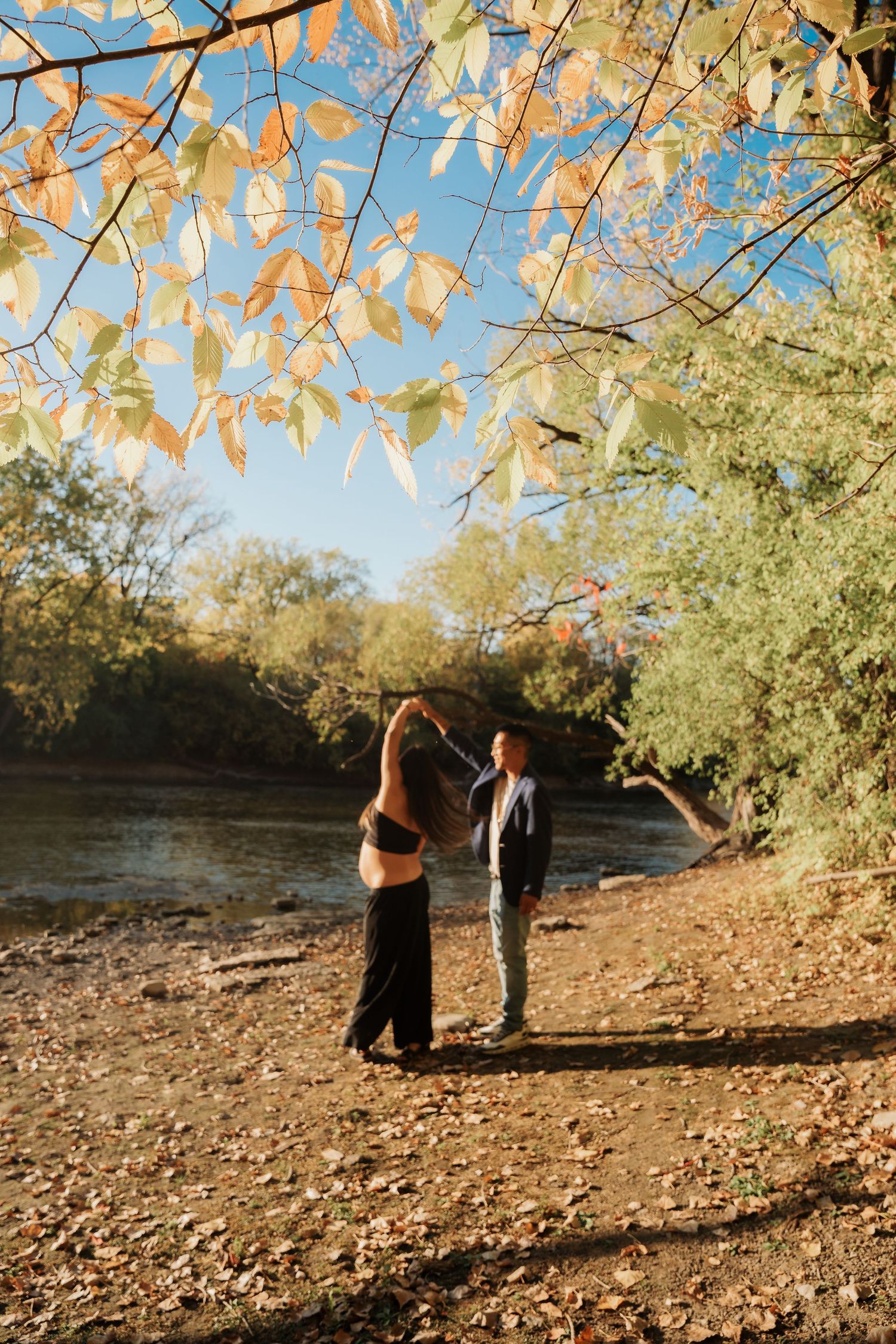 A pregnant couple holds hands near a sunlit river surrounded by colorful autumn foliage.