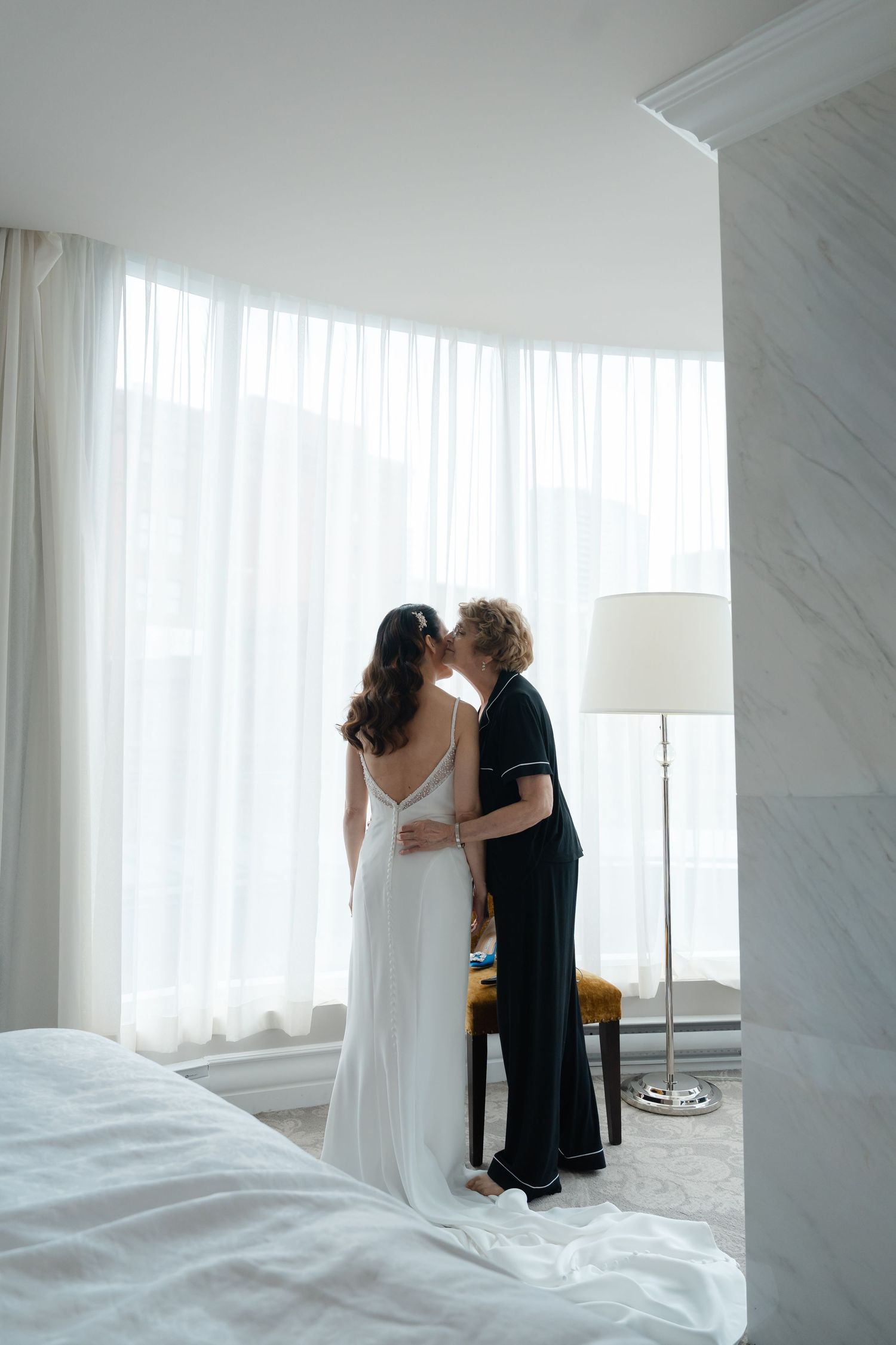 A bride in a white gown shares a romantic kiss with a partner in a bright, elegant hotel room.