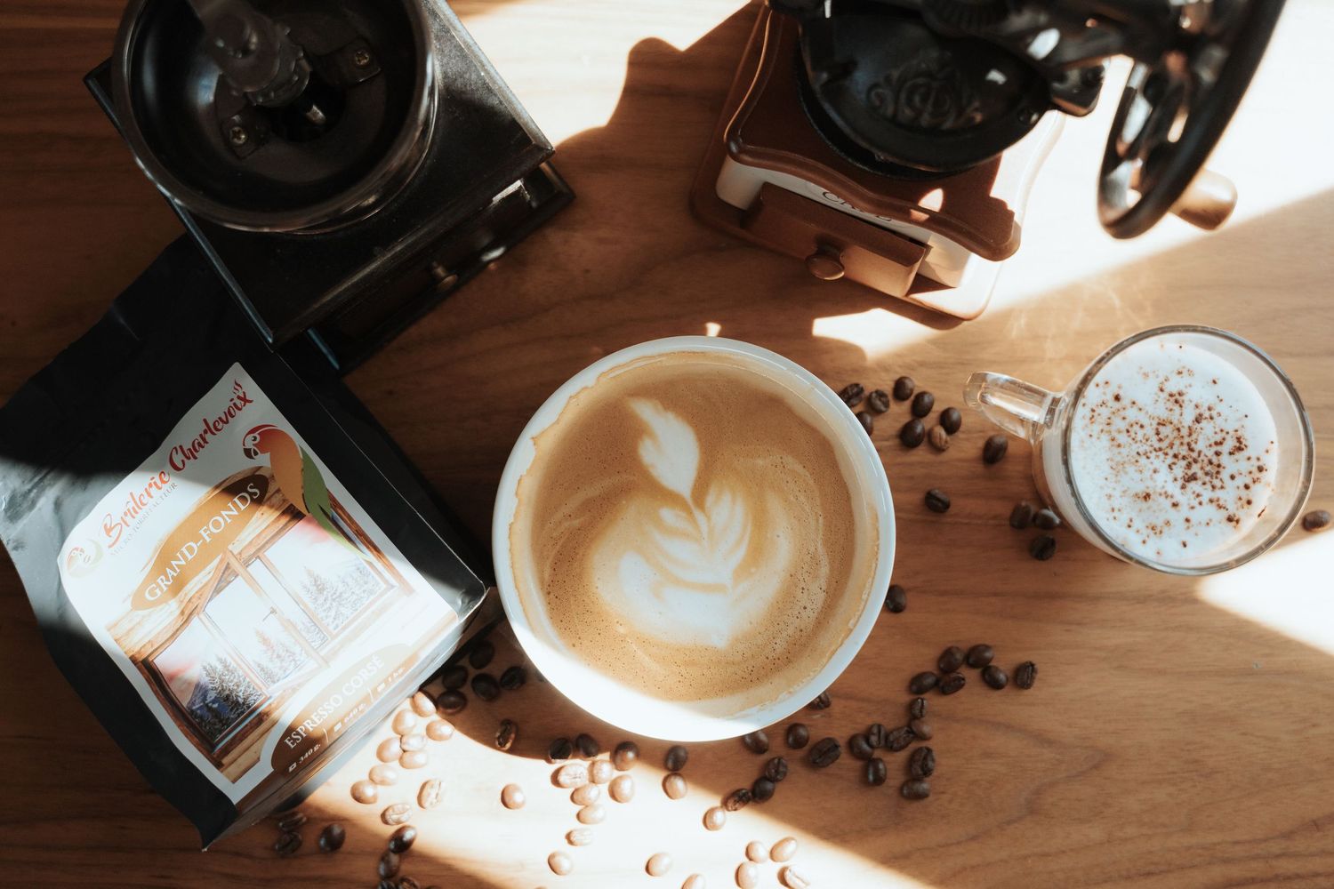 Overhead view of latte art coffee cup with coffee beans, grinder, and coffee bag on wooden table.