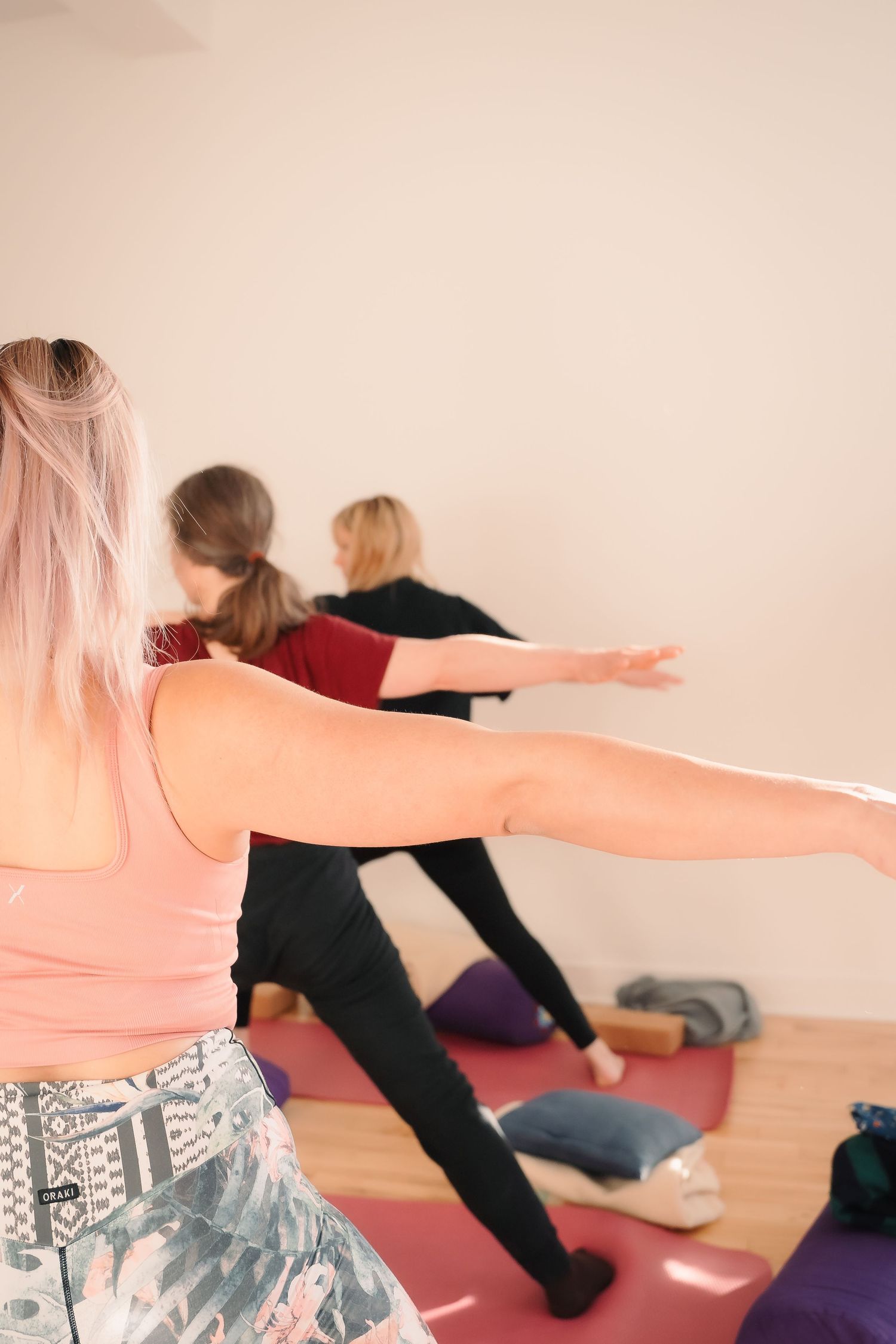Three women practicing warrior pose with arms extended during a yoga class in a bright studio.