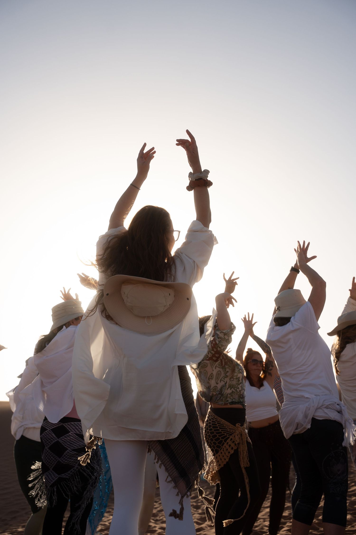 Silhouette of group raising hands toward glowing sunset sky on sandy desert dunes.