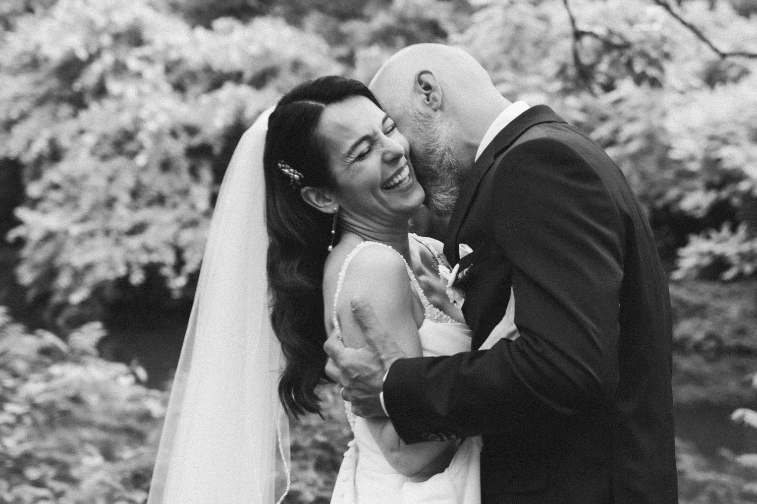 Black and white photo of a laughing bride in veil and groom sharing a joyful intimate moment outdoors.