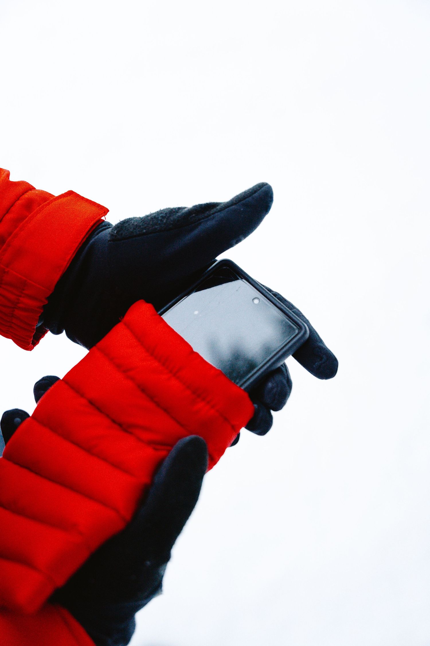 Hands in black gloves and red winter jacket holding a smartphone outdoors in cold weather.