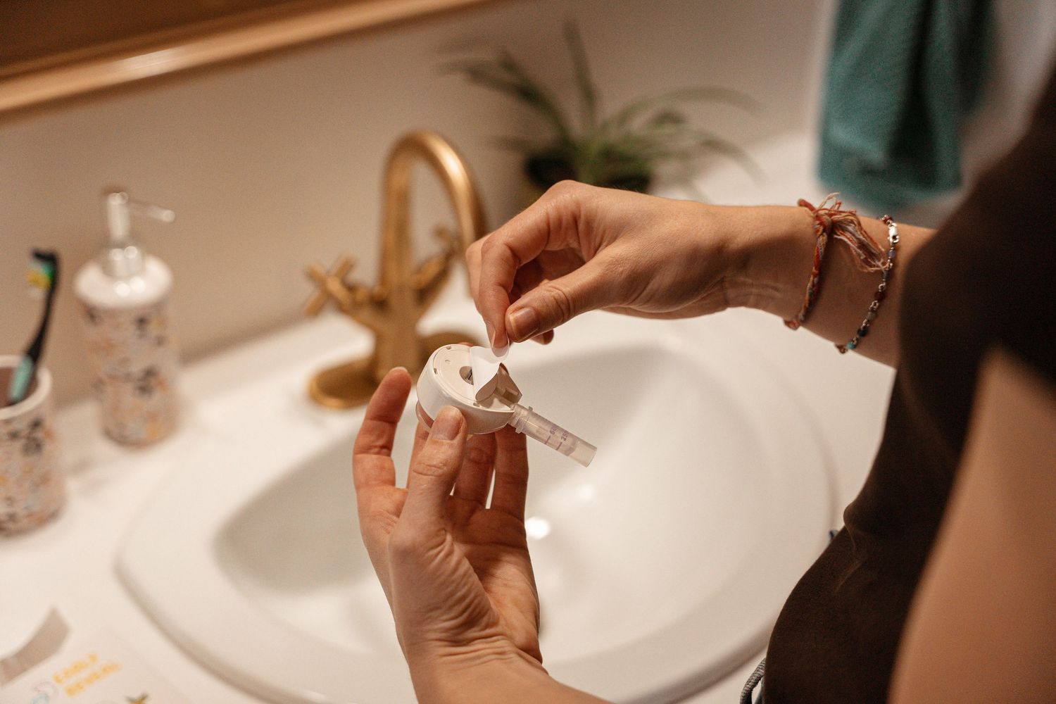 Person unwrapping a small medical device over a bathroom sink with gold fixtures.