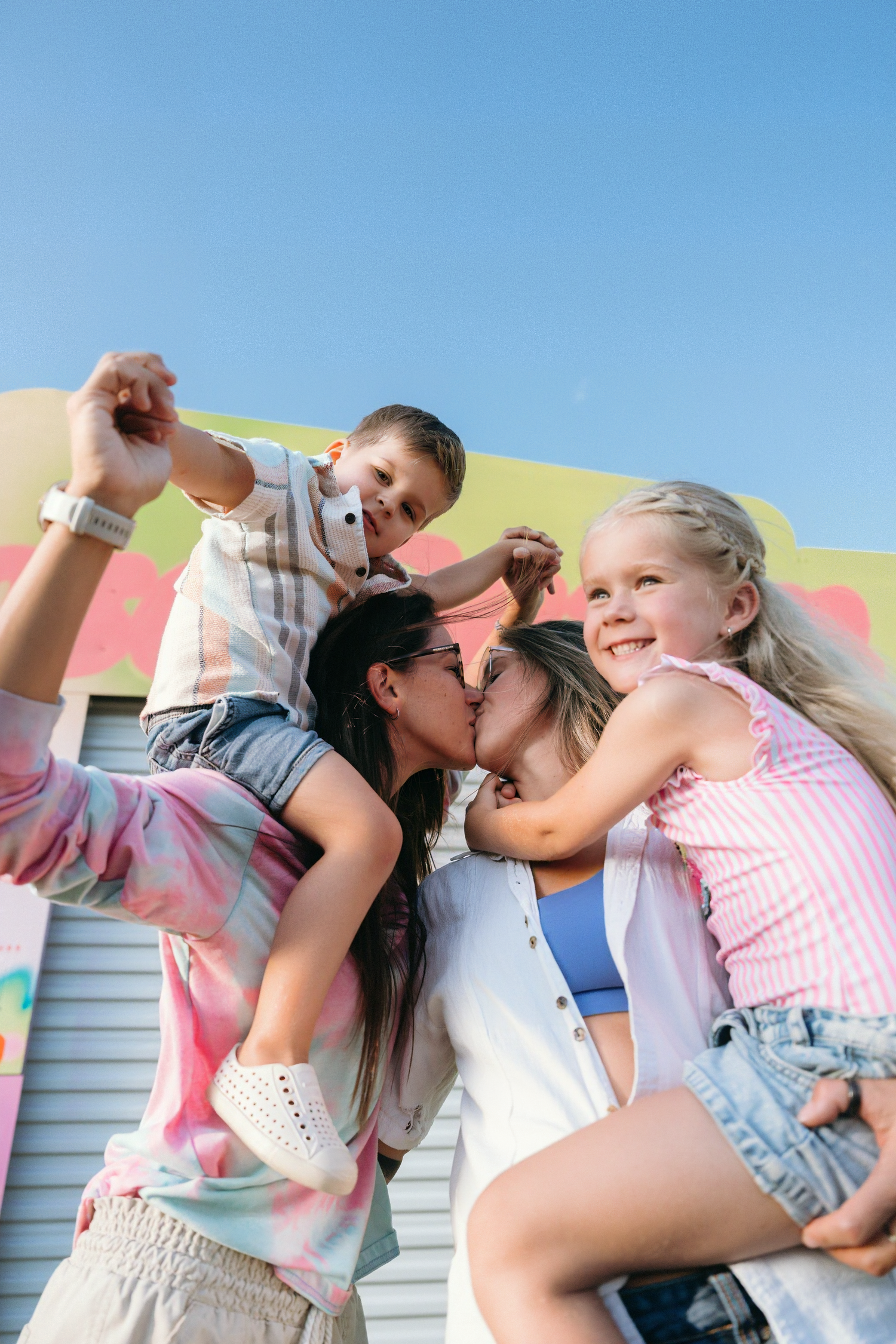 Two women kissing while giving children piggyback rides outdoors on a sunny day near a colorful building.