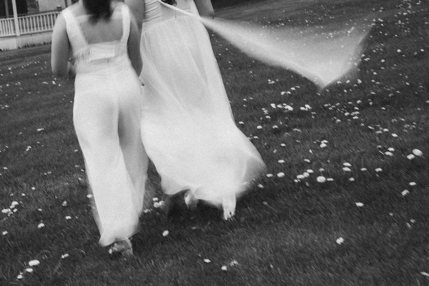 Black and white photo of two brides in white gowns walking across a flower-strewn lawn.