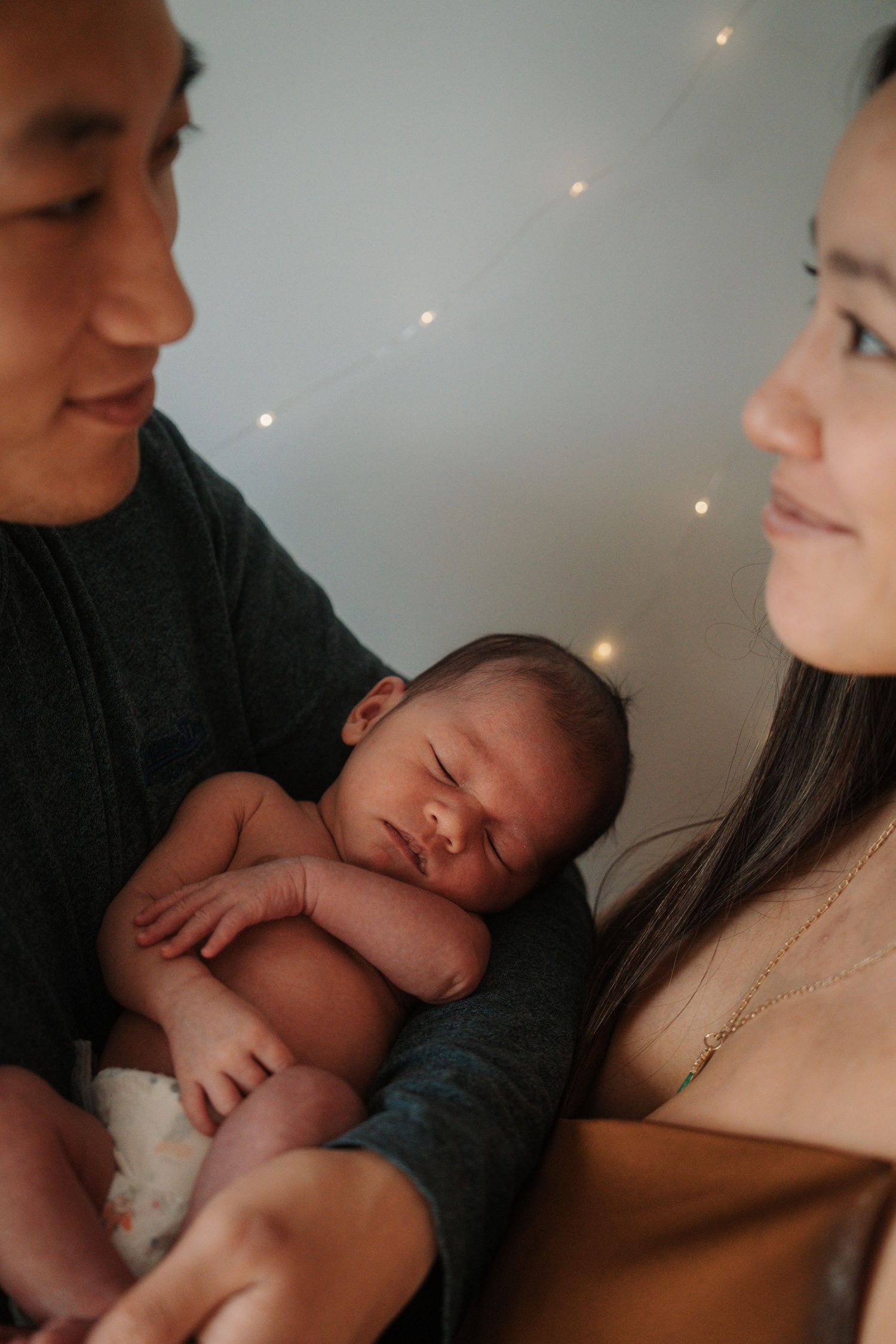 New parents smiling lovingly at sleeping newborn baby with fairy lights behind.