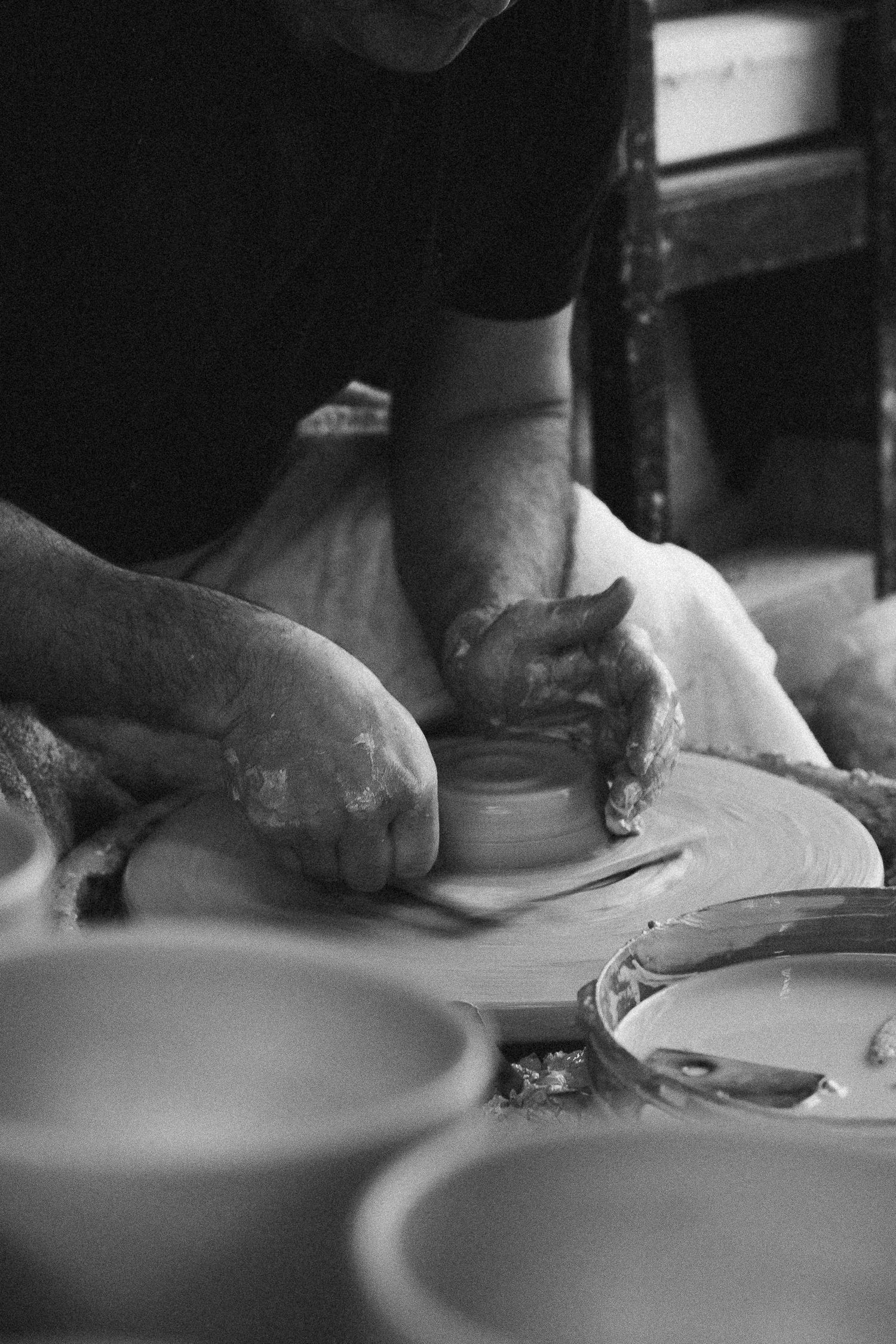 Black and white photo of a potter shaping clay on a wheel, hands covered in slip, with ceramic bowls nearby.