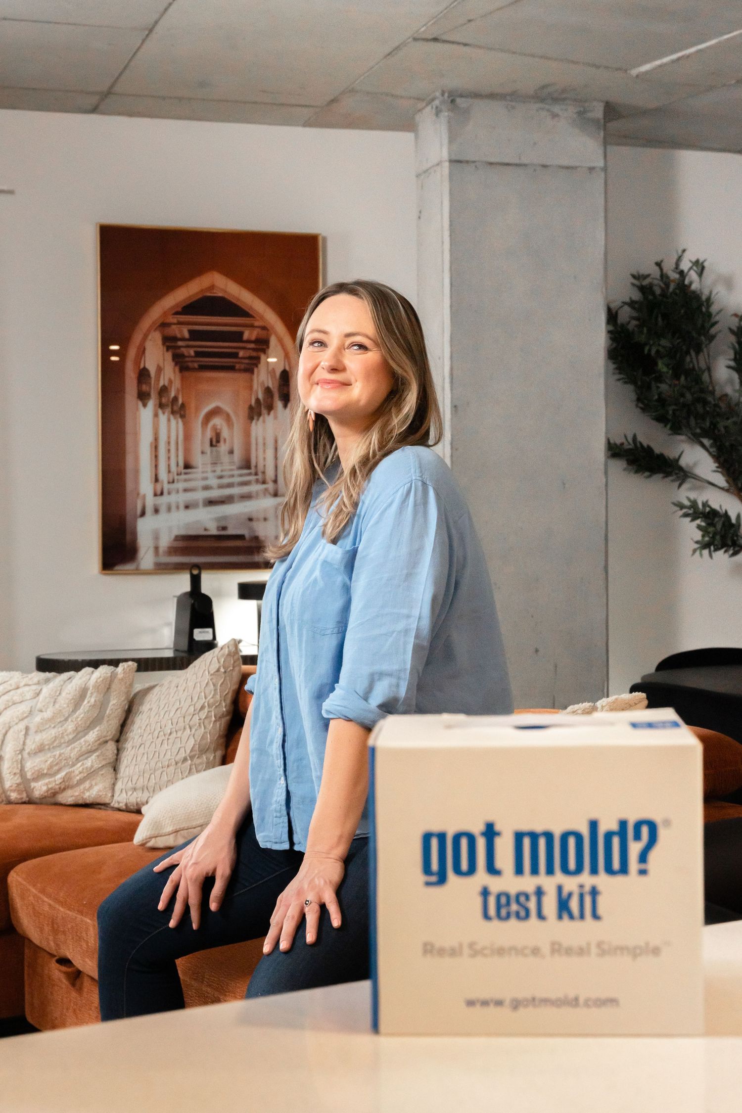 Smiling woman leaning against a table beside a Got Mold test kit box in a modern living space.