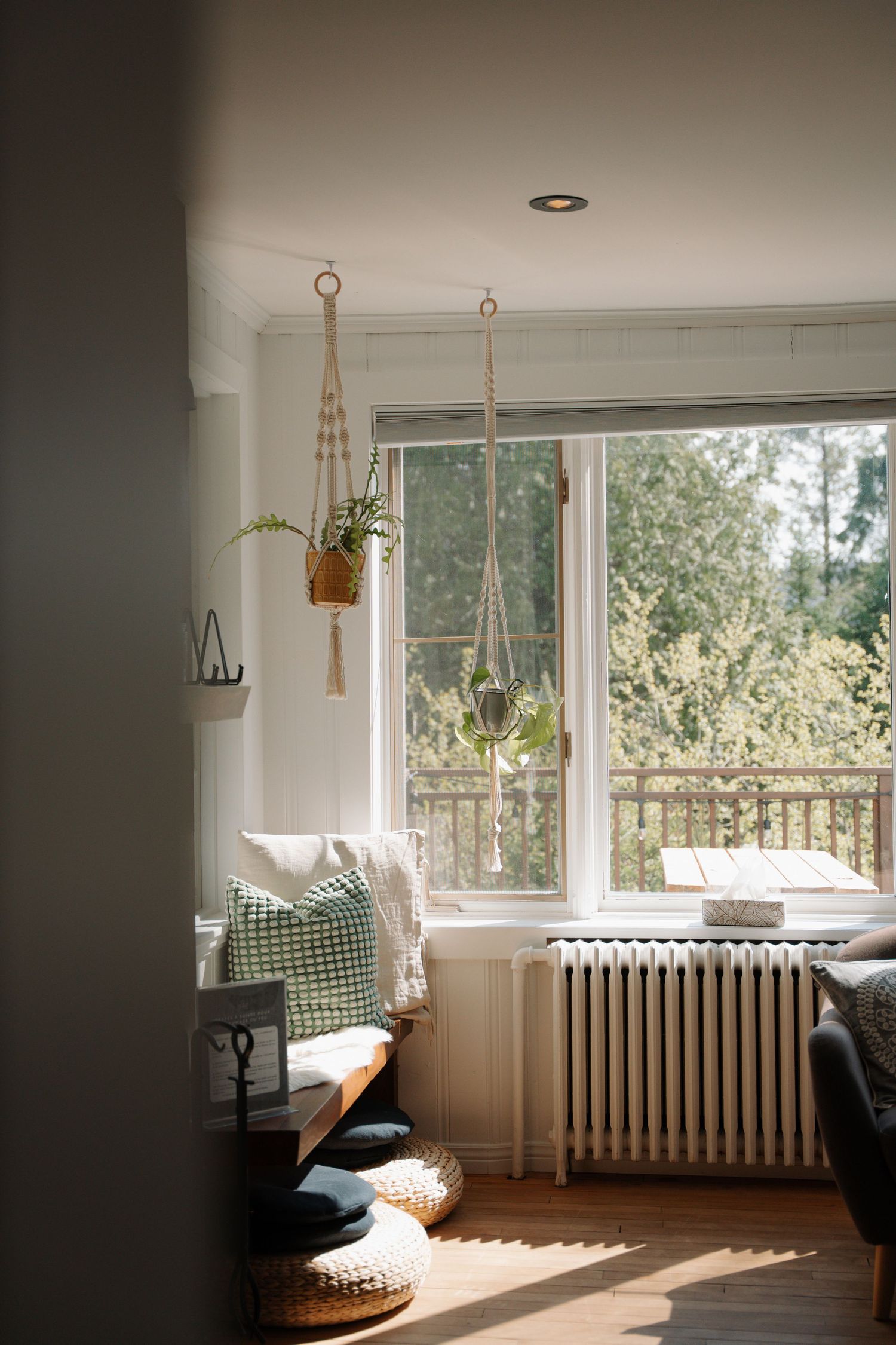 Bright living room corner with macrame plant hangers, green cushions, radiator, and large window overlooking lush trees.