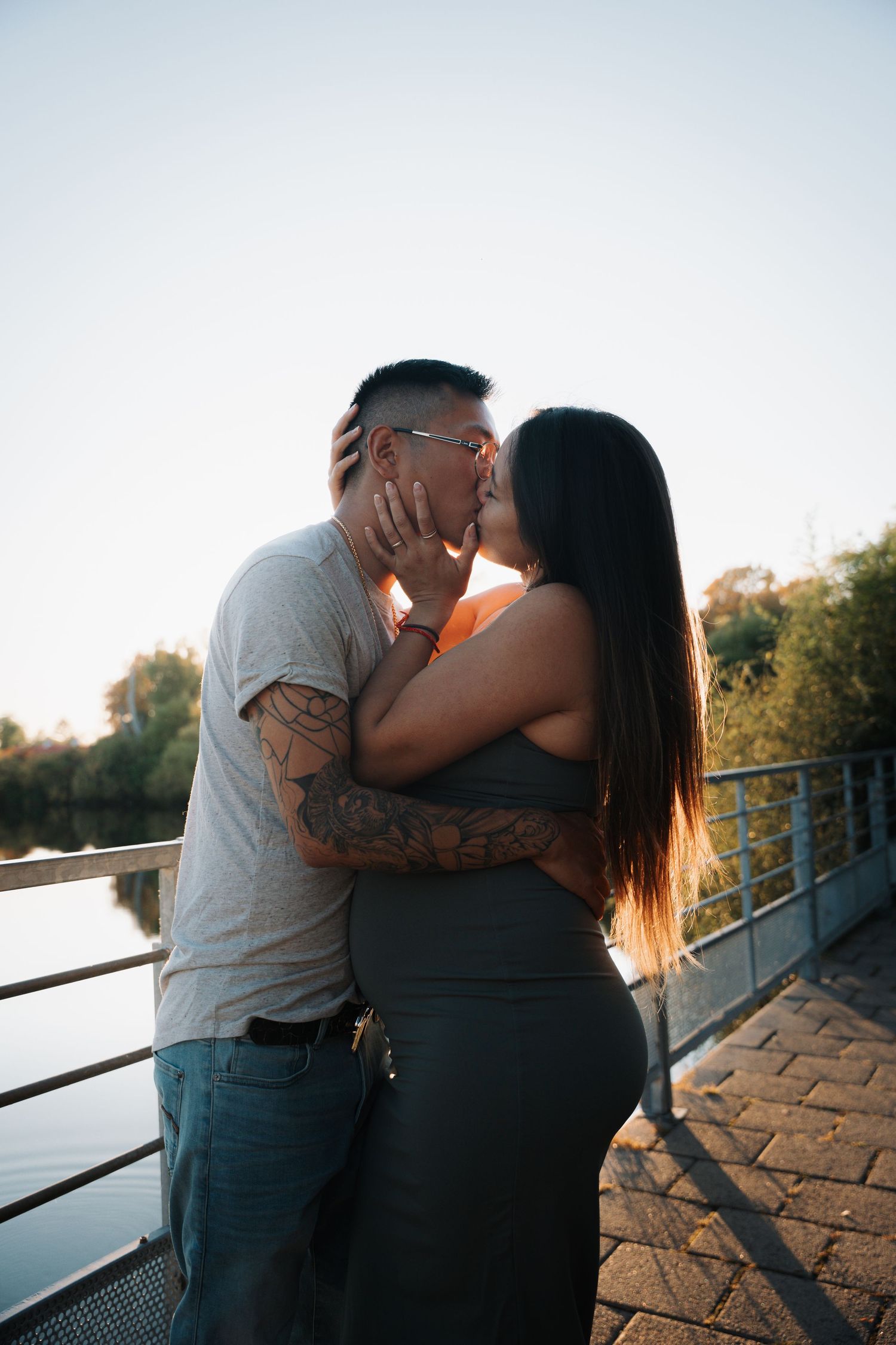 Tattooed couple kissing on a waterfront bridge at sunset with trees and water visible in the background.