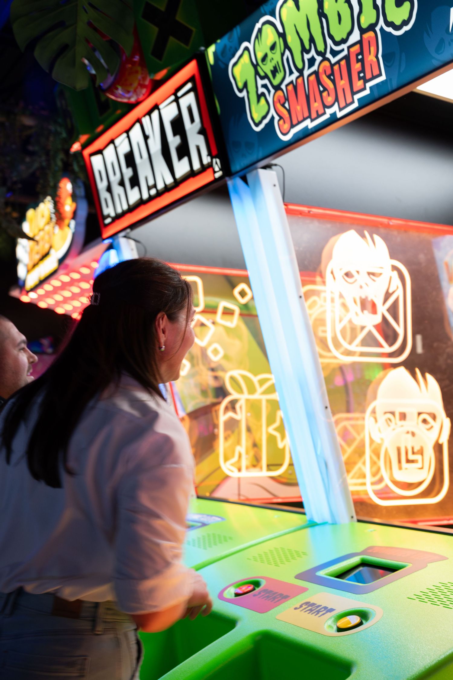 Woman plays Zombie Smasher arcade game with glowing neon zombie targets at an arcade.