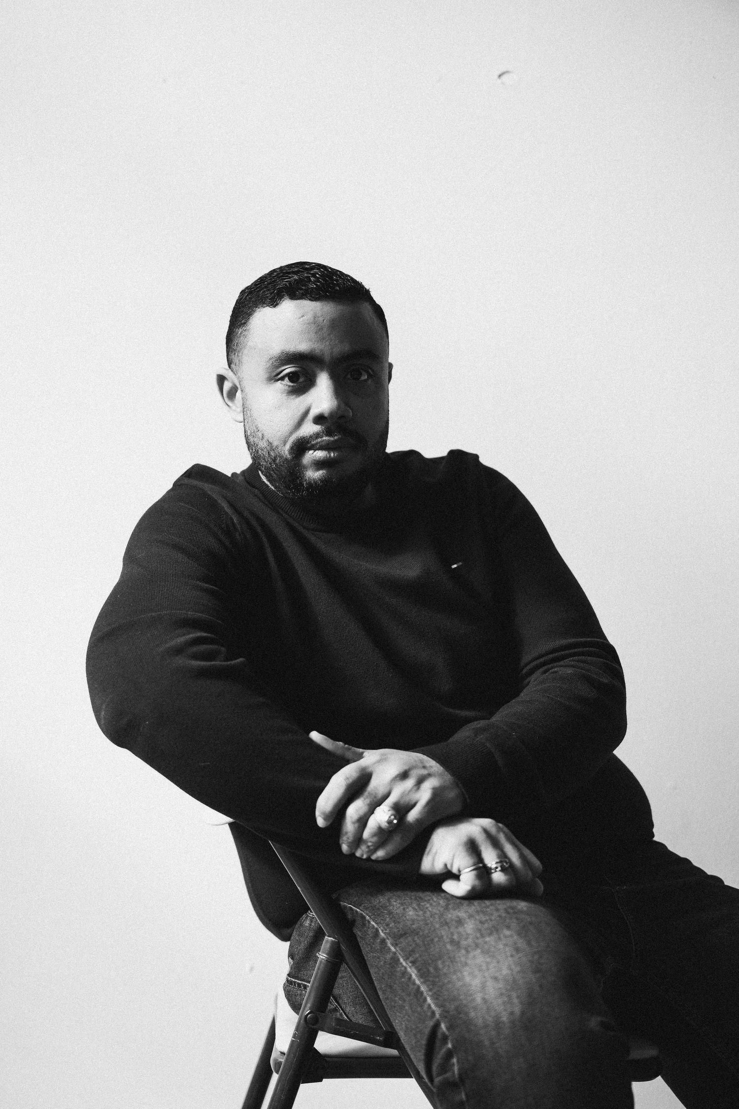 Black and white portrait of a bearded man in a black sweatshirt sitting on a chair against a white background.