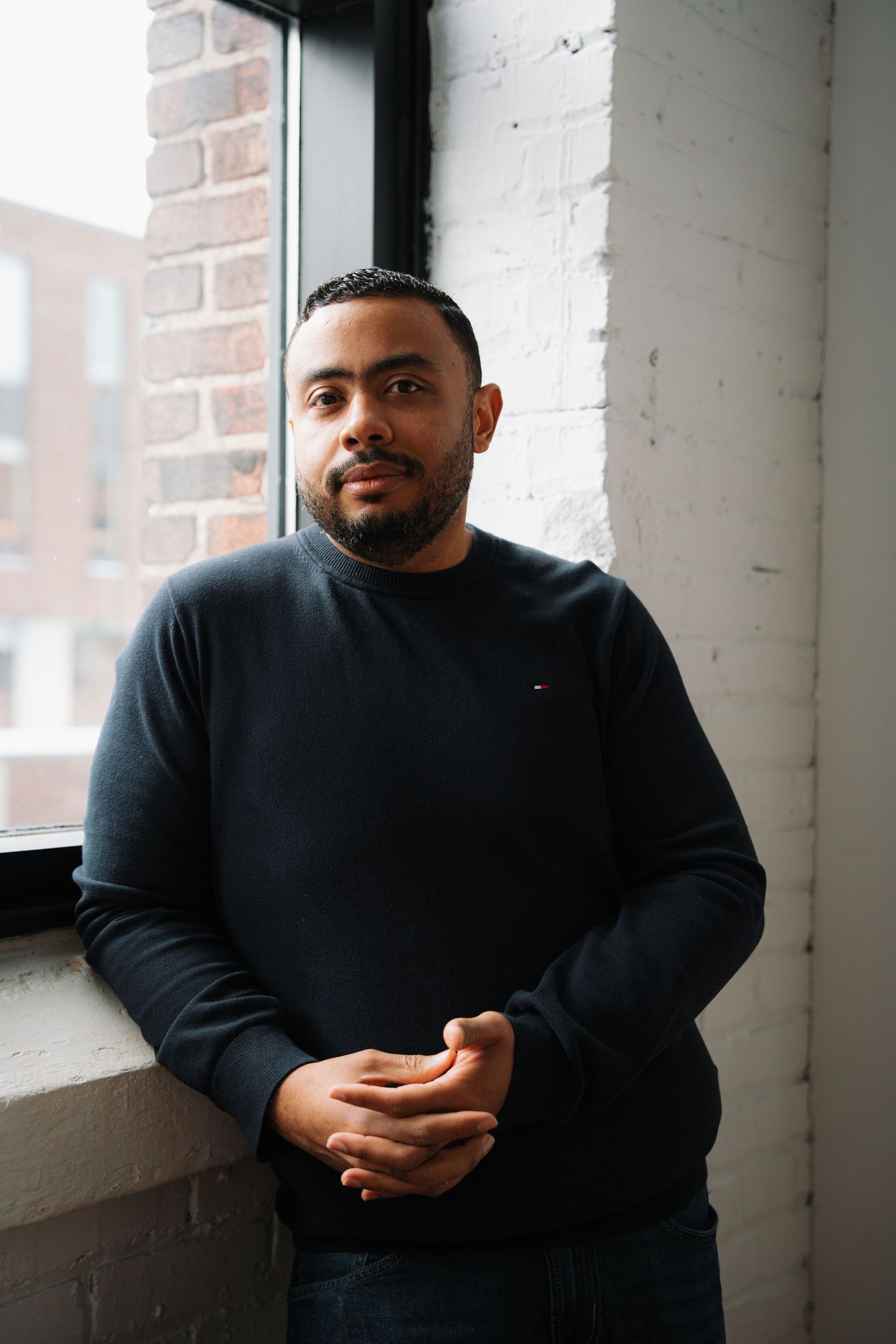Man in black Tommy Hilfiger sweater standing by window with brick wall background.