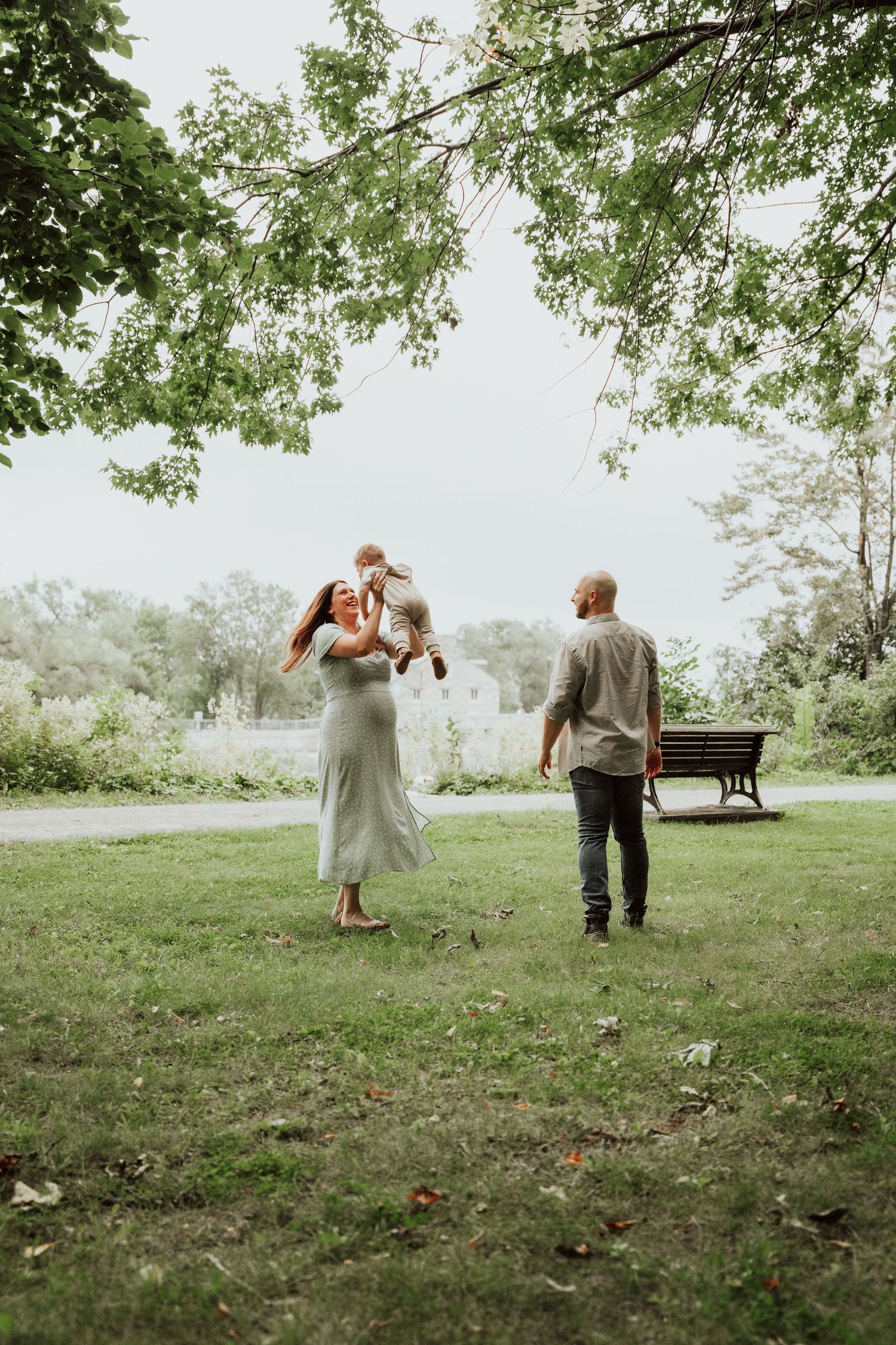 Pregnant woman in mint dress lifts baby while partner walks toward them in green park.