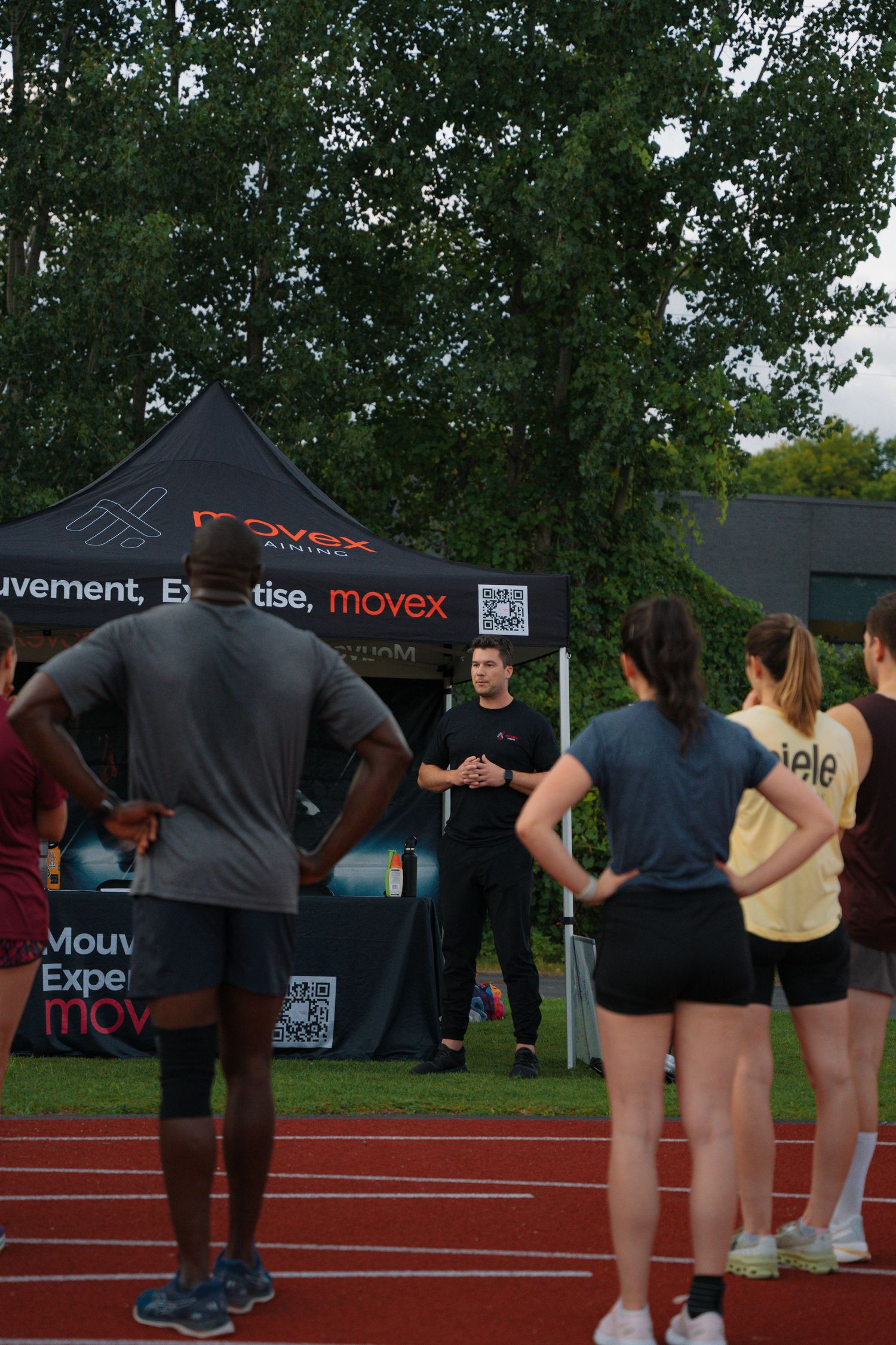 Coach in black addressing a group of athletes on a track near a Movex branded tent.
