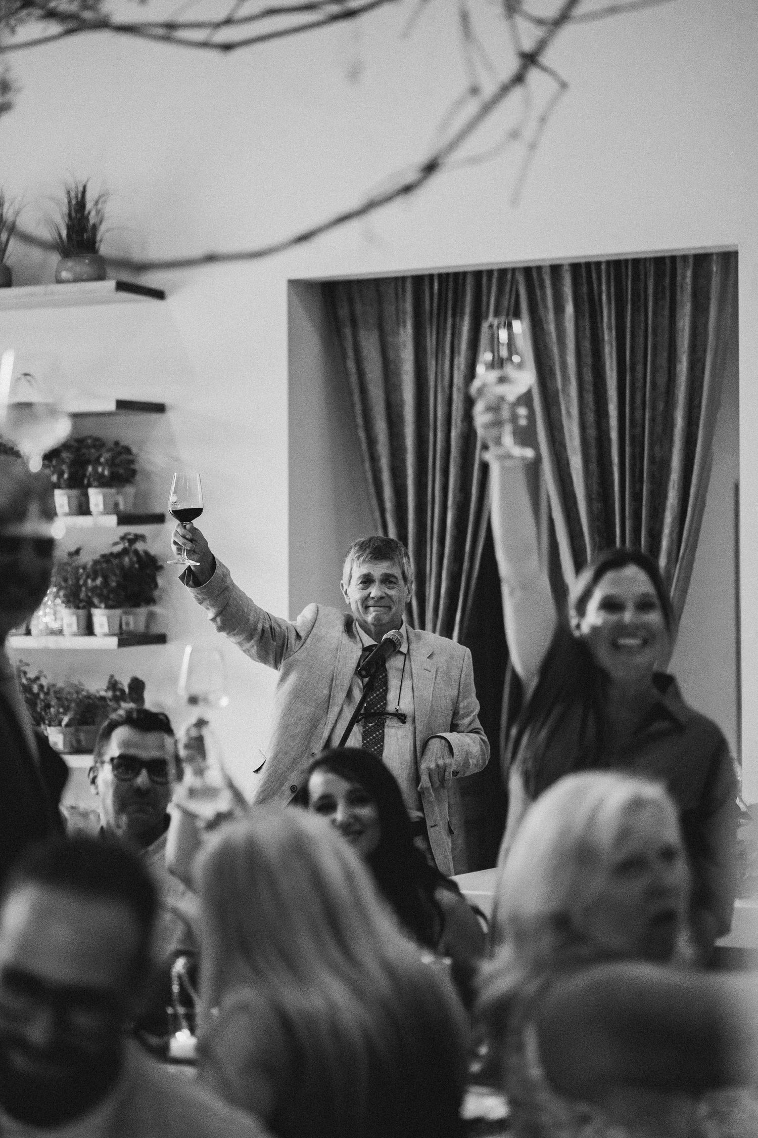Black and white photo of a man raising a wine glass in a toast at a wedding reception.