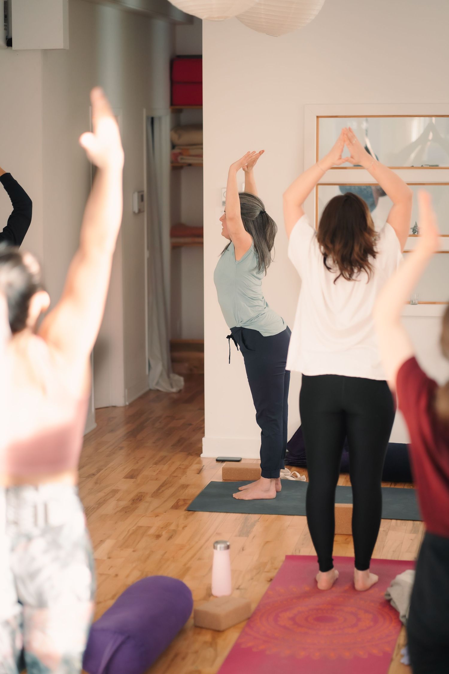 Group of yoga students performing a standing pose with arms raised overhead in a bright studio.