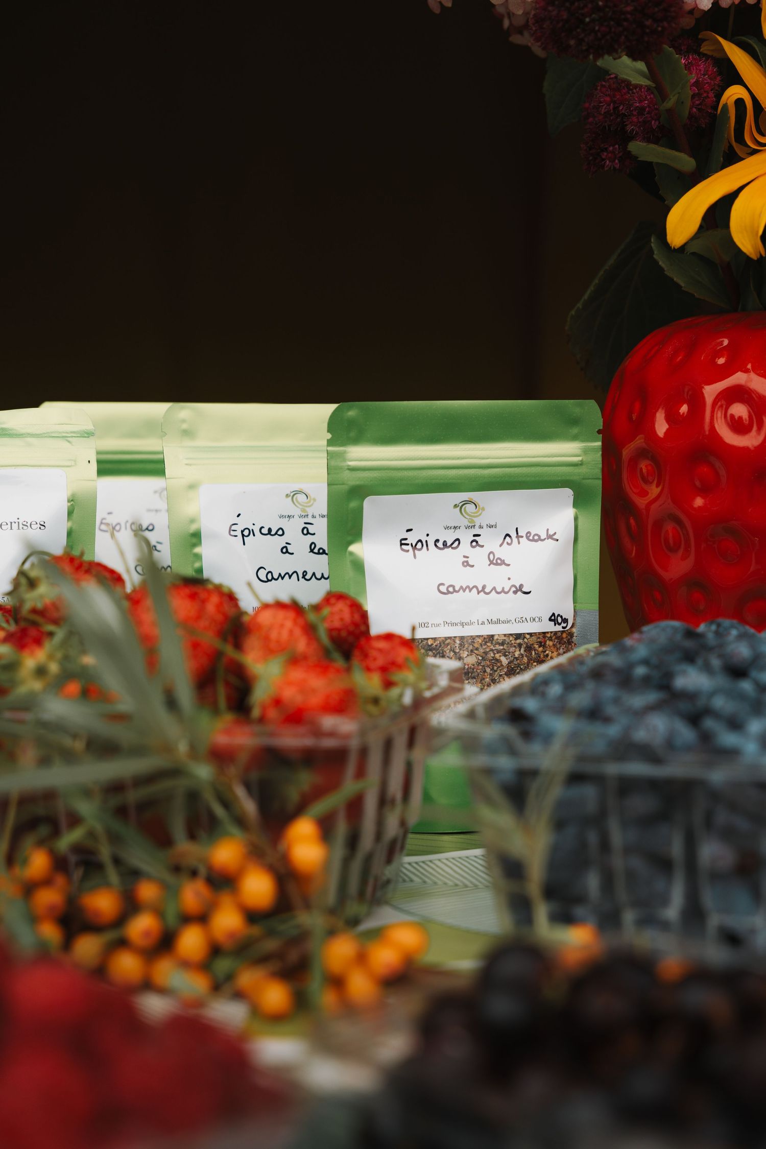 Green packaged spice blends with handwritten labels displayed beside fresh berries at a market.