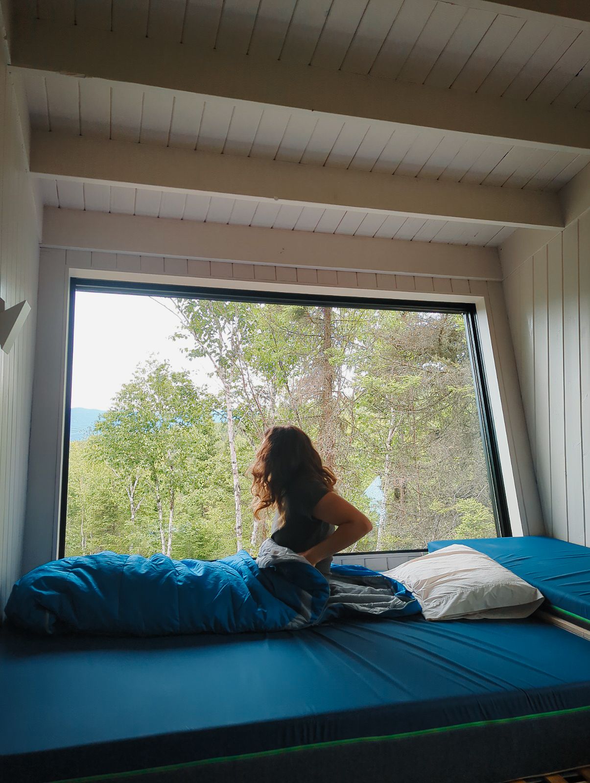 Woman sitting on bed gazing through large cabin window at lush green forest and lake view.