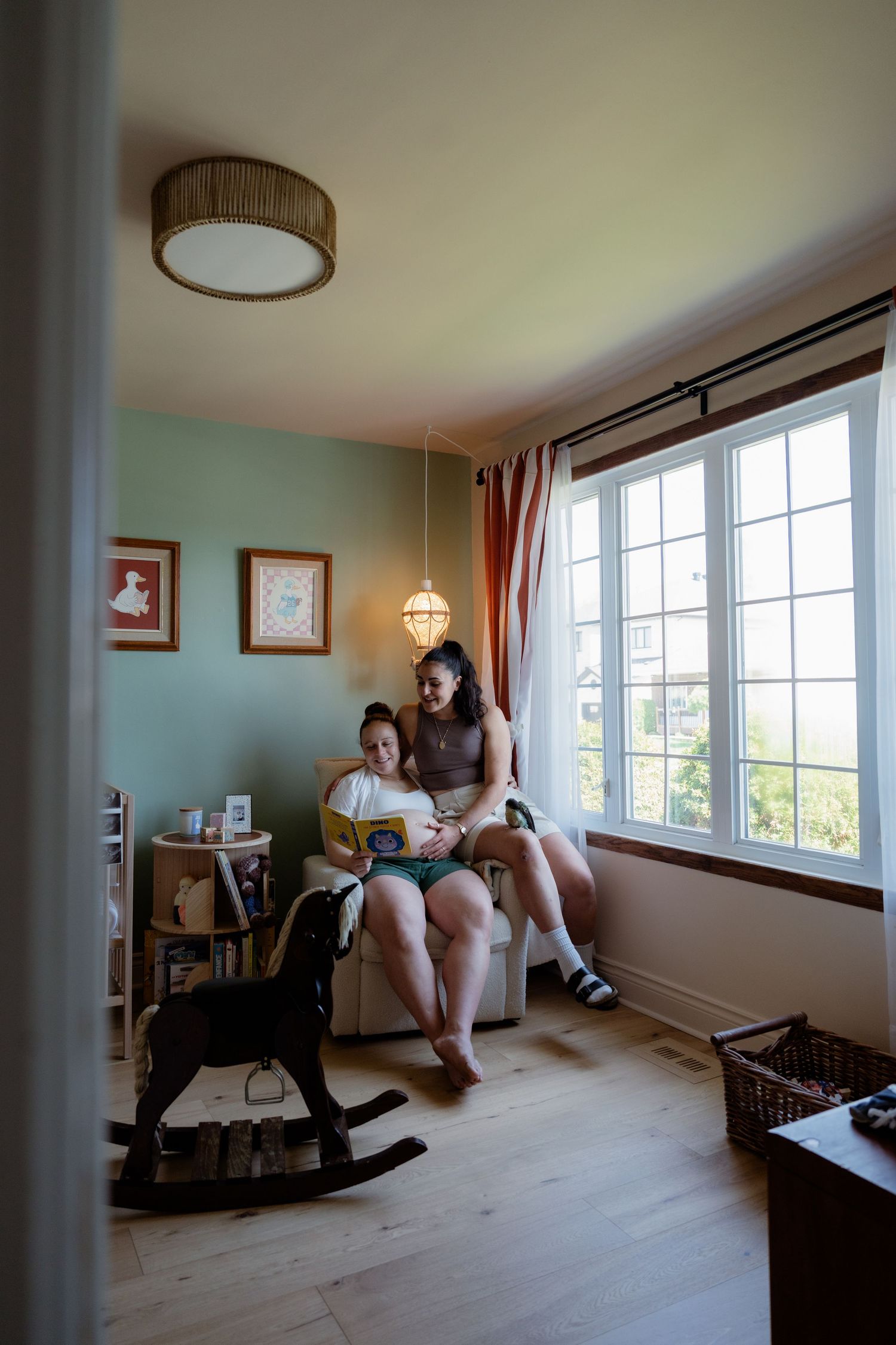 A pregnant woman and her partner sit together in a cozy nursery reading a book by a bright window.