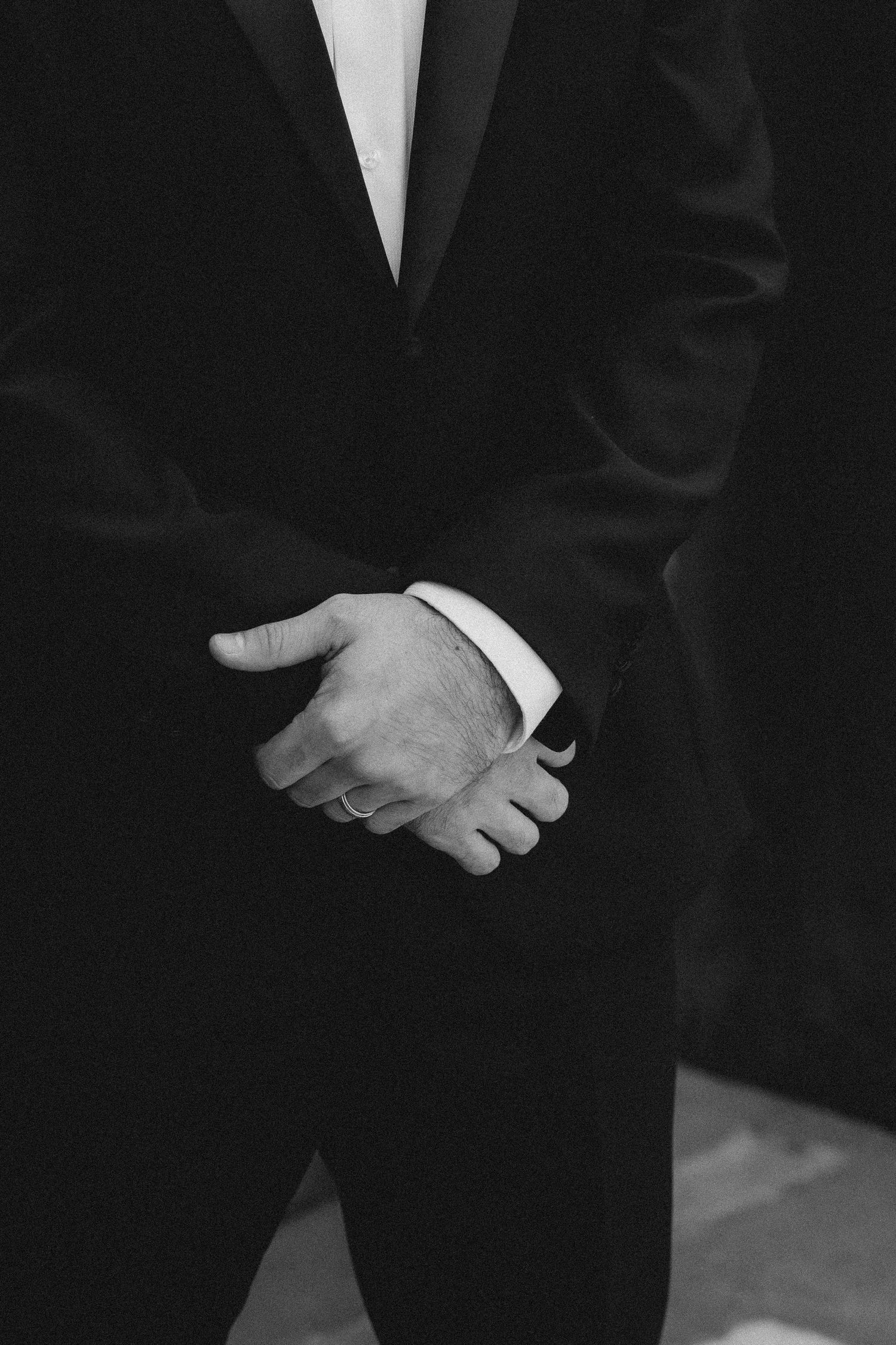 Black and white close-up of groom in tuxedo clasping hands behind his back.