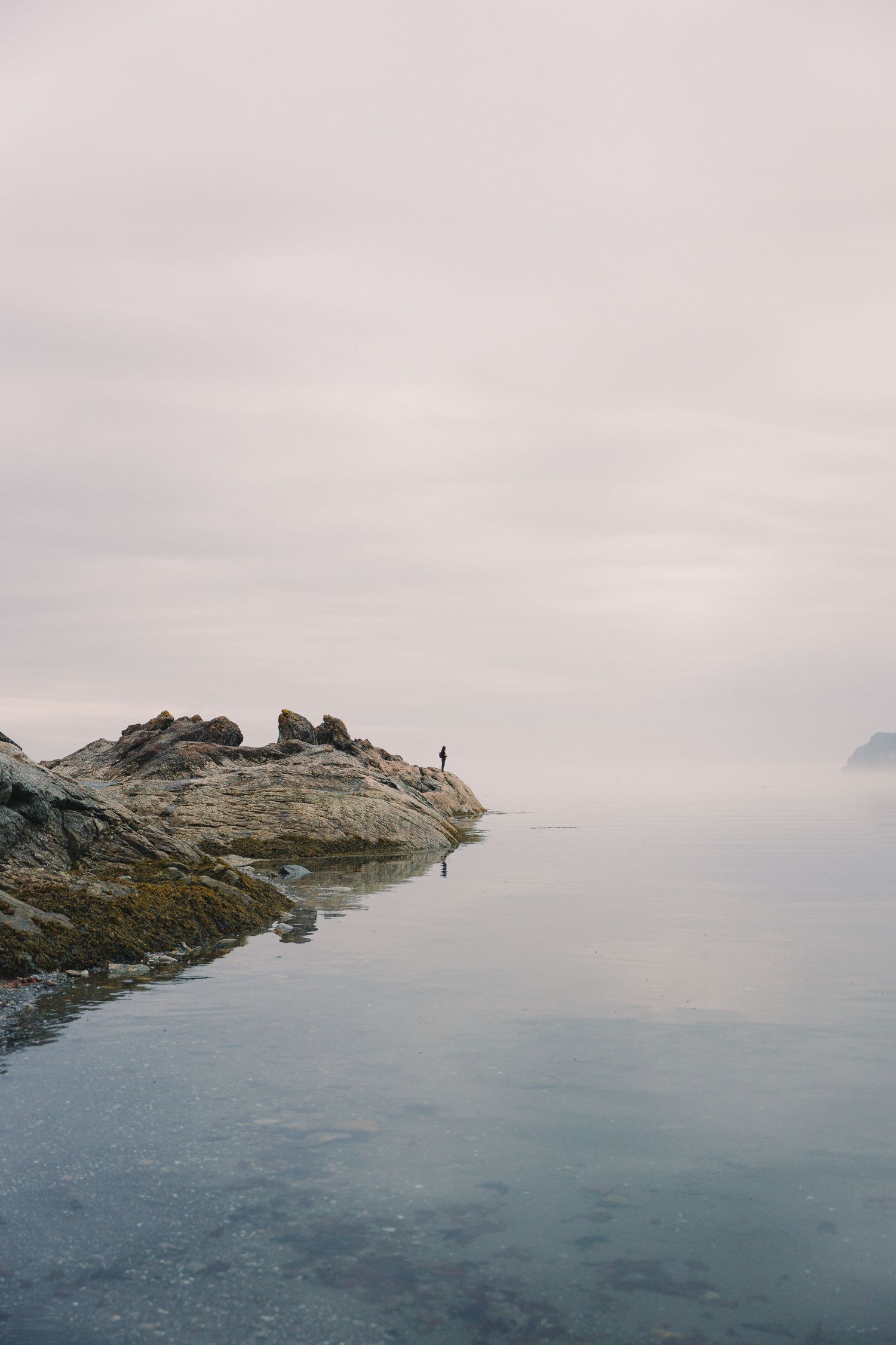 A solitary figure stands on misty rocky shoreline reflected in calm still ocean waters on an overcast day.