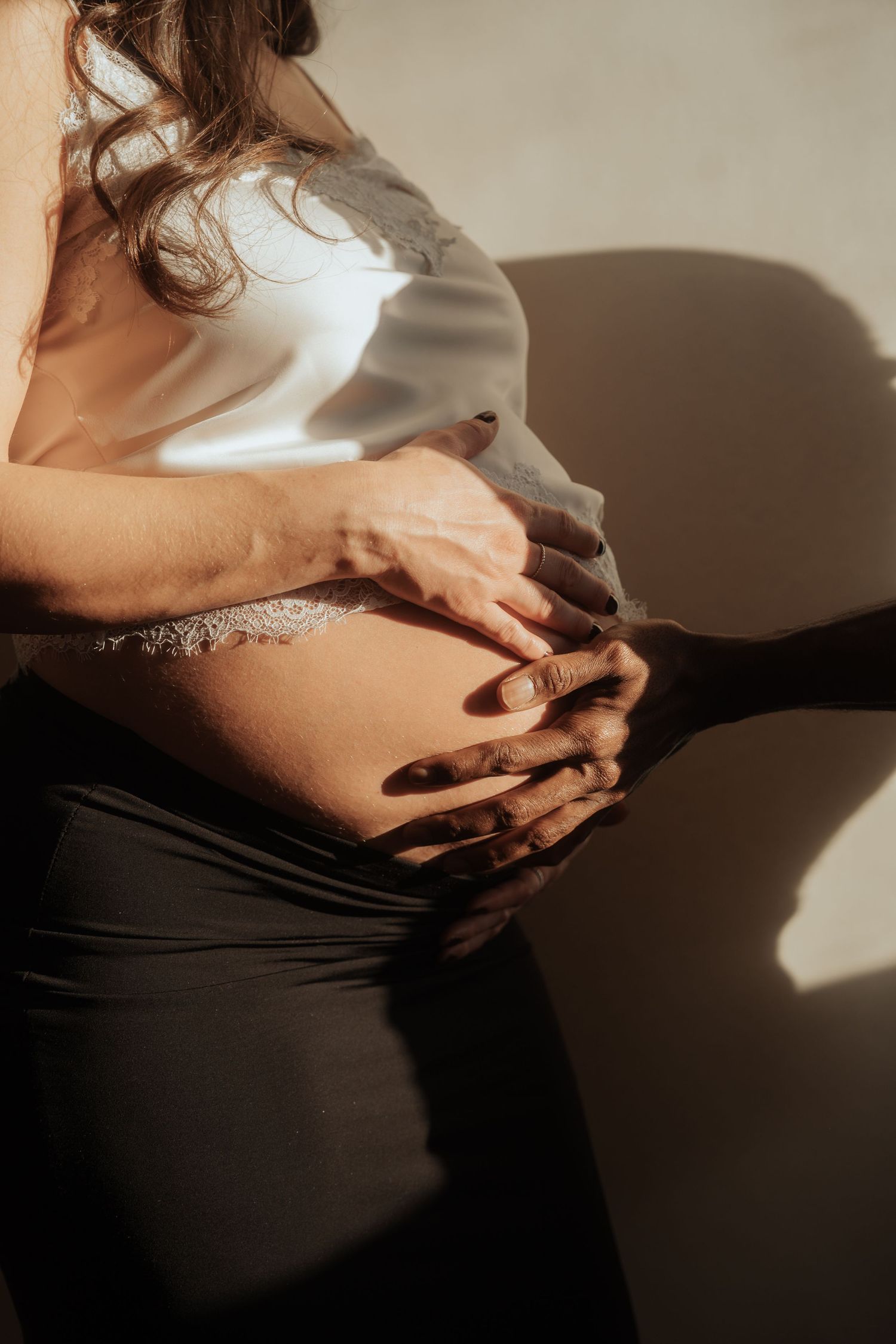 A couple tenderly holding a pregnant belly together in warm sunlight casting soft shadows on a neutral wall.