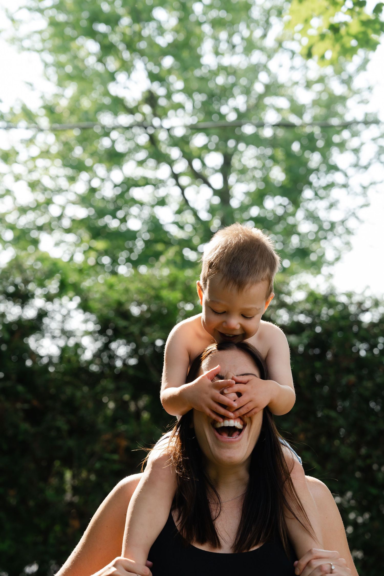 A laughing mother carries a happy toddler on her shoulders outdoors, with lush green trees in the background.